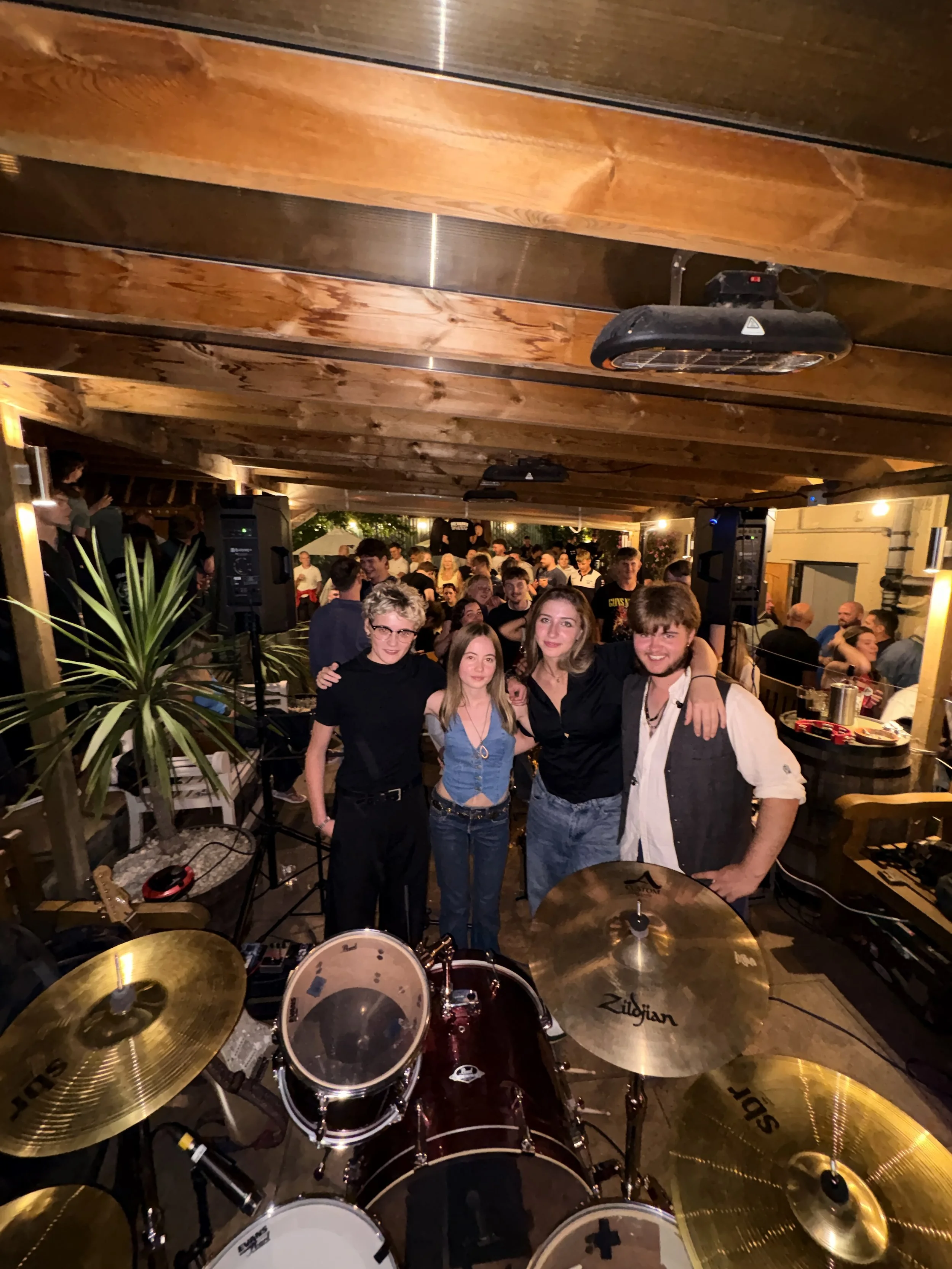 Four people standing close together, smiling, with a crowded indoor event in the background, a drum set in the foreground.