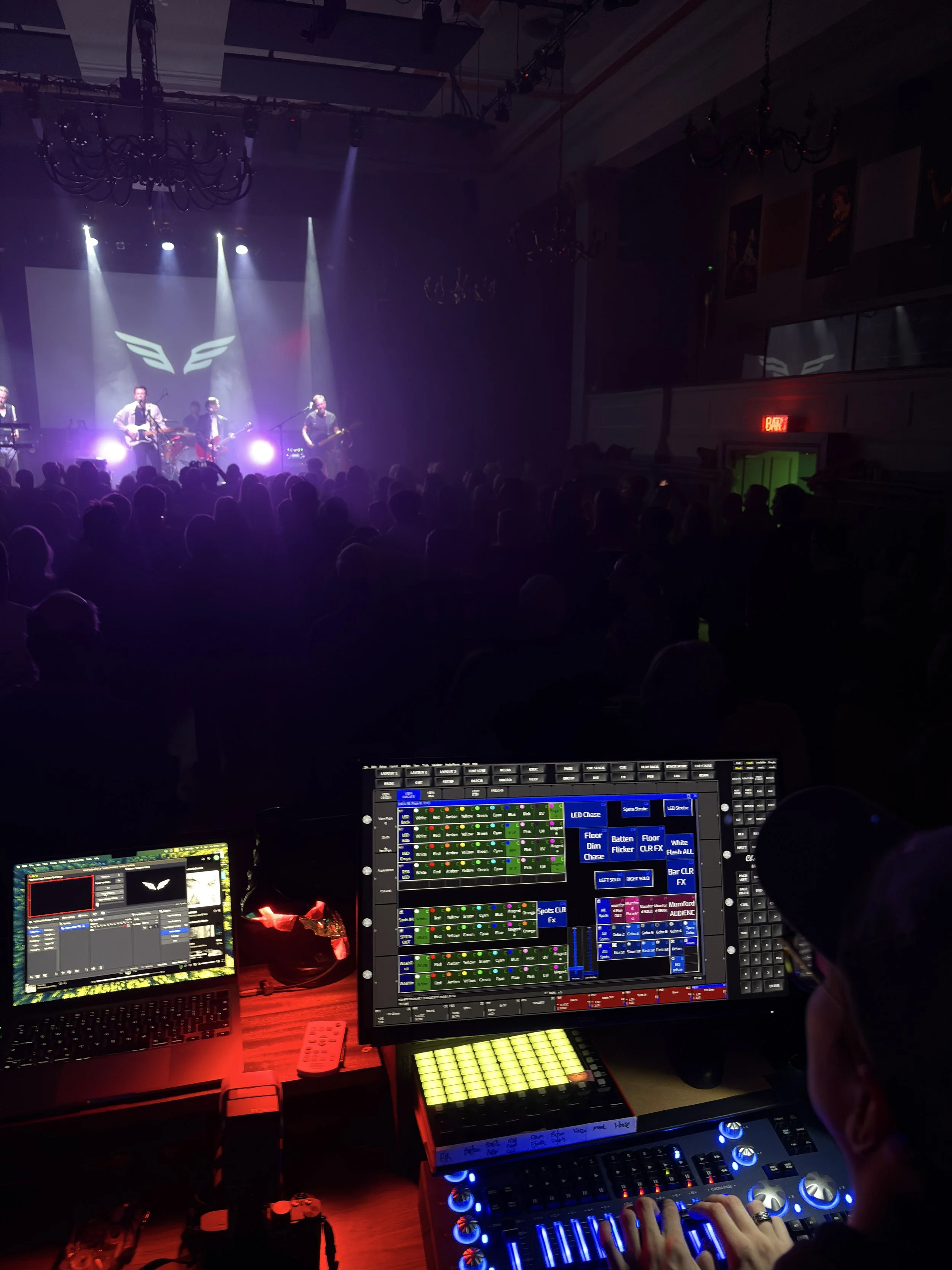 Stage with band performing, audience in the dark, illuminated by spotlights, in a concert hall. Mixing consoles with lights and controls are in foreground.