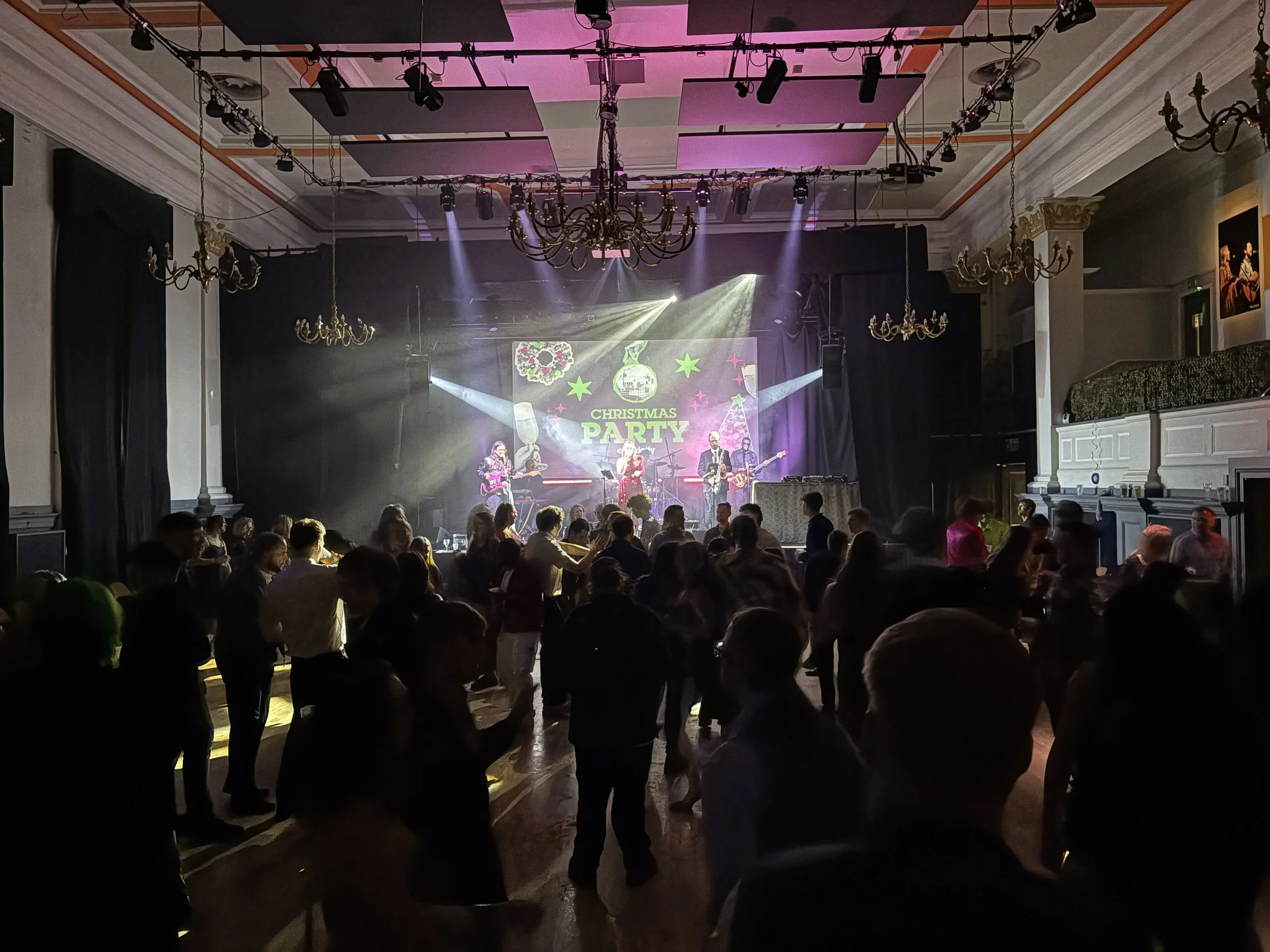 People dancing and mingling at a Christmas party with live band performing on stage, holiday lights, and a large screen displaying Christmas-themed graphics in the background.