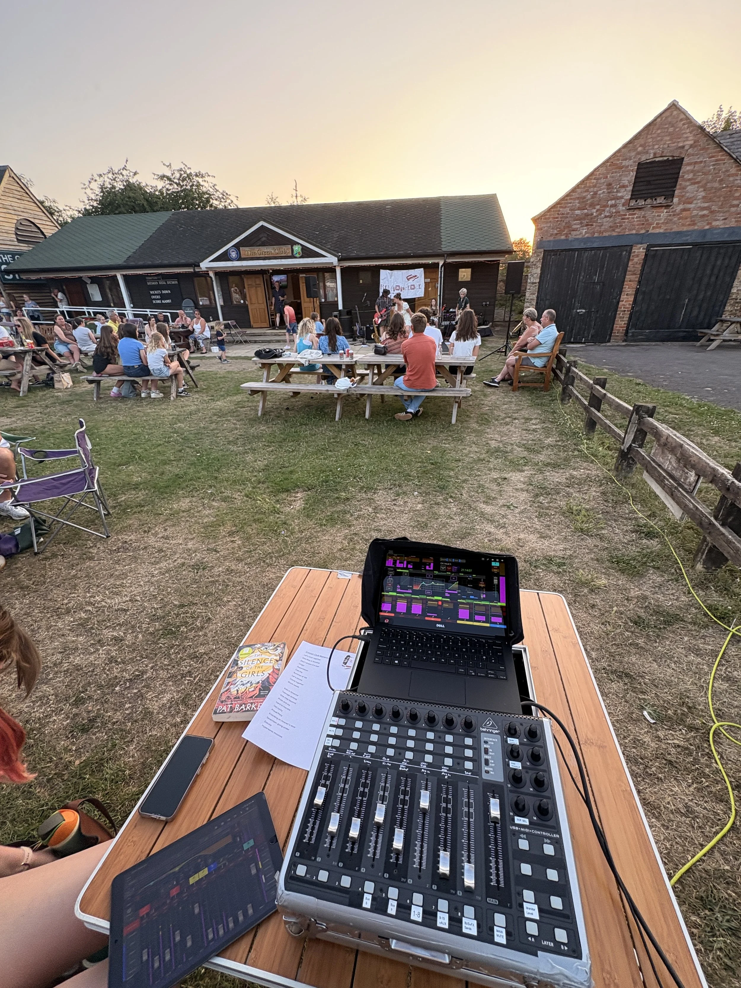 Outdoor concert at a venue with a band performing on stage, audience seated at picnic tables, and a sound mixer and electronic devices on a wooden table in the foreground.