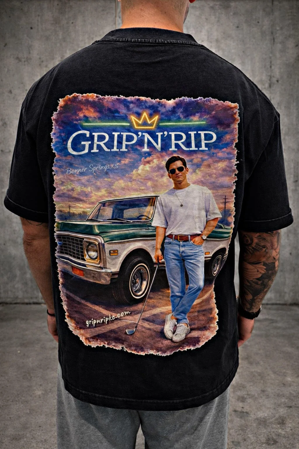 Back of a person wearing a black T-shirt with a large graphic print. The graphic depicts a man in sunglasses standing next to a vintage car, holding a golf club. The background shows a colorful sky with clouds. Text on the shirt reads "GRIP'N'RIP" with a yellow crown above the text, and a website URL is at the bottom.