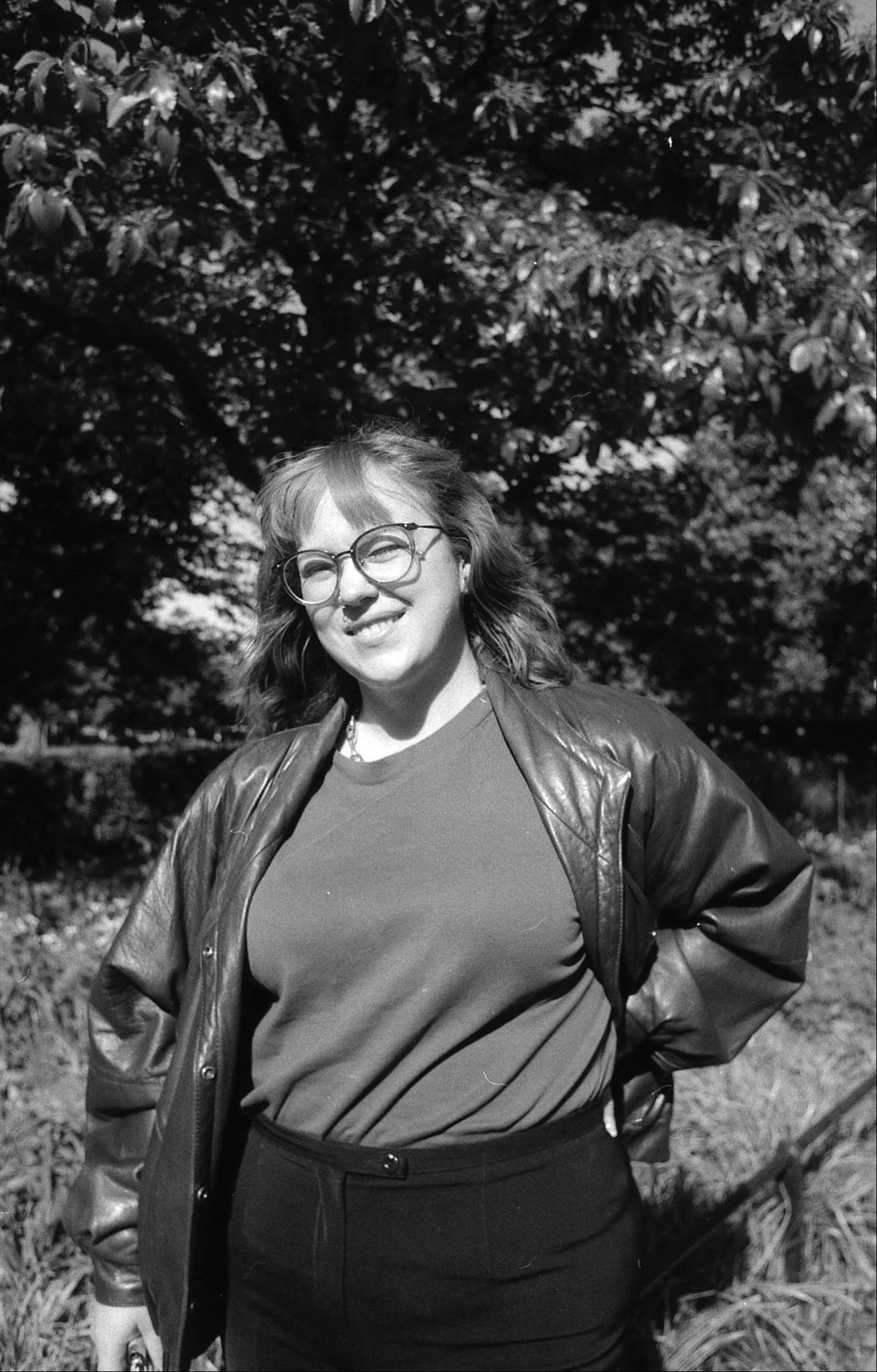 A woman with wavy hair and glasses smiling outdoors, wearing a leather jacket over a shirt, standing in front of trees.