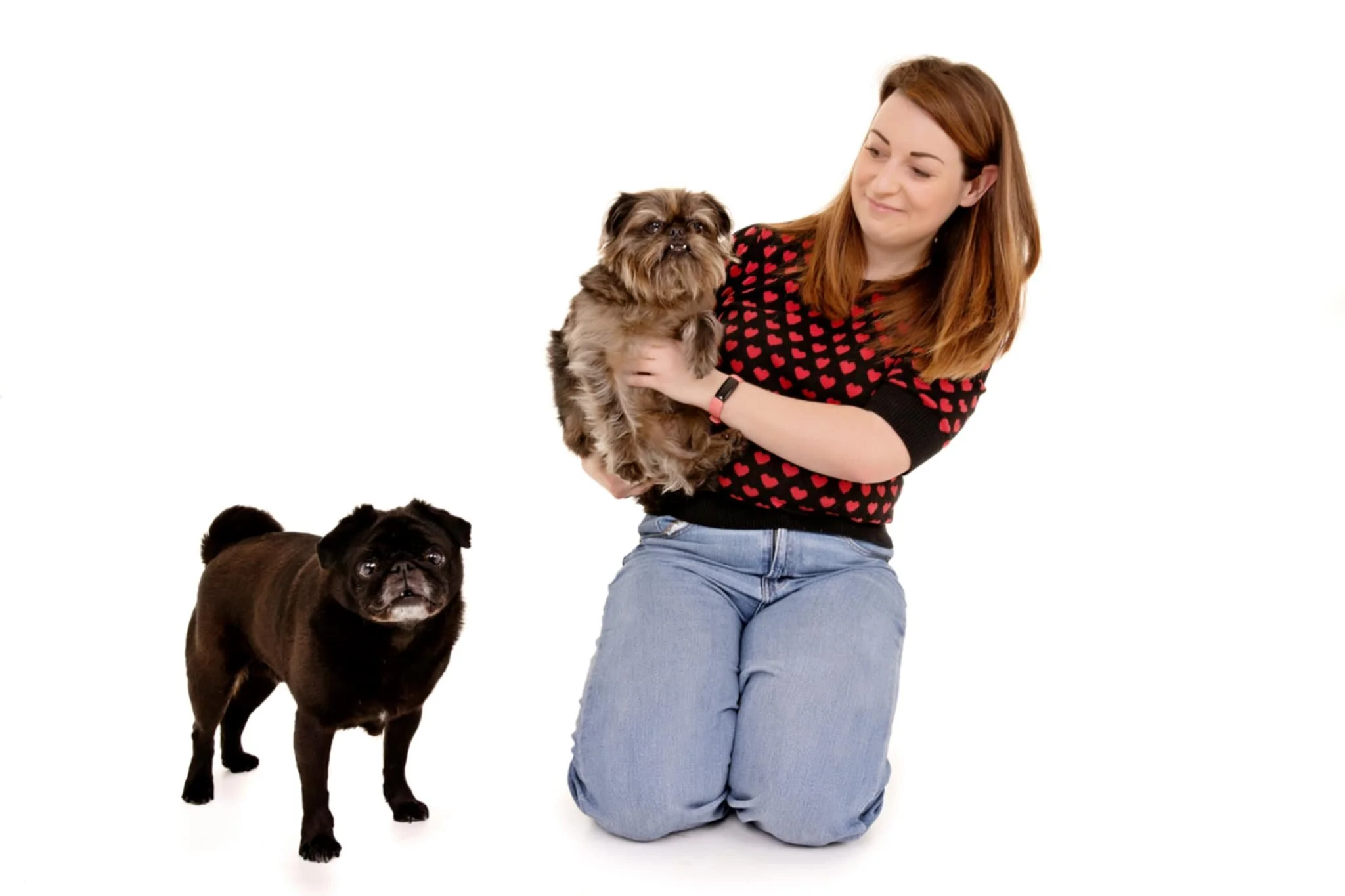A woman kneeling on the floor, holding a small brown and black dog with a fluffy coat while another black pug stands next to her, looking up.