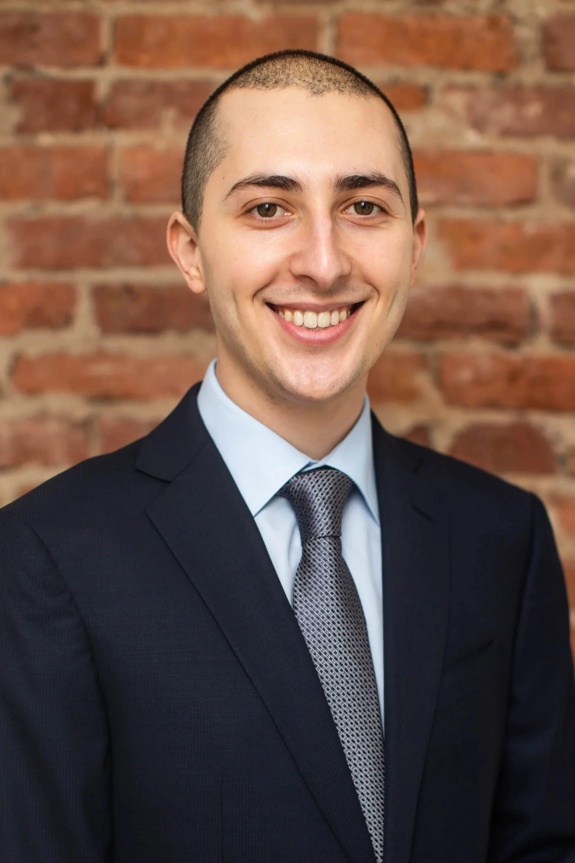 Portrait of a smiling man in a navy suit, light blue shirt, and patterned tie, standing in front of a brick wall.