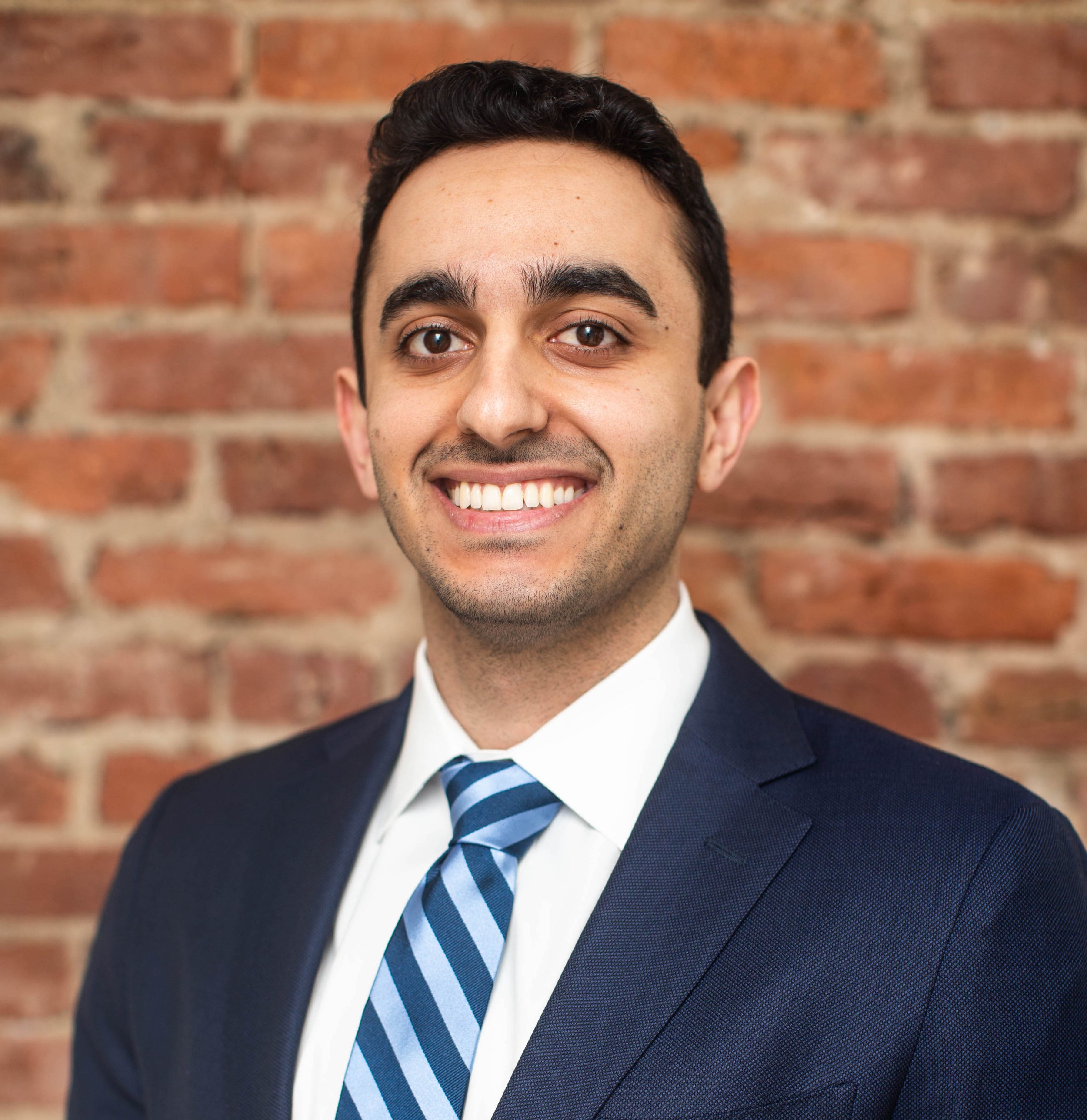 Professional headshot of a smiling man in a suit with a brick wall background.