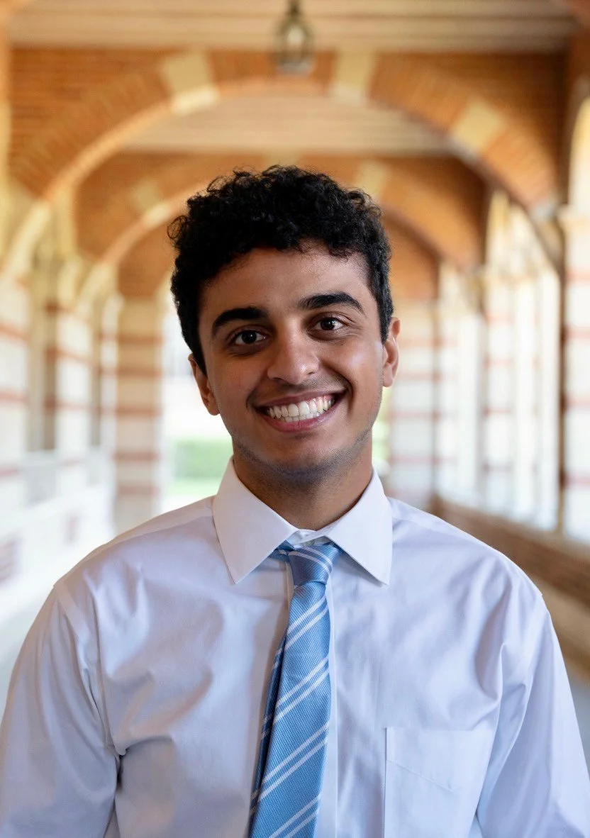 A young man with dark curly hair and a bright smile, wearing a white shirt and a light blue striped tie, standing in a hallway with brick archways and large windows.