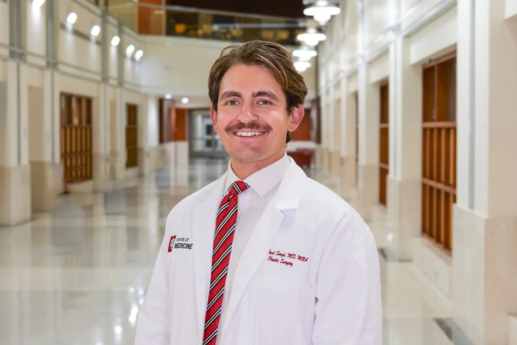 A smiling male doctor in a white coat and red striped tie standing in a hospital corridor.