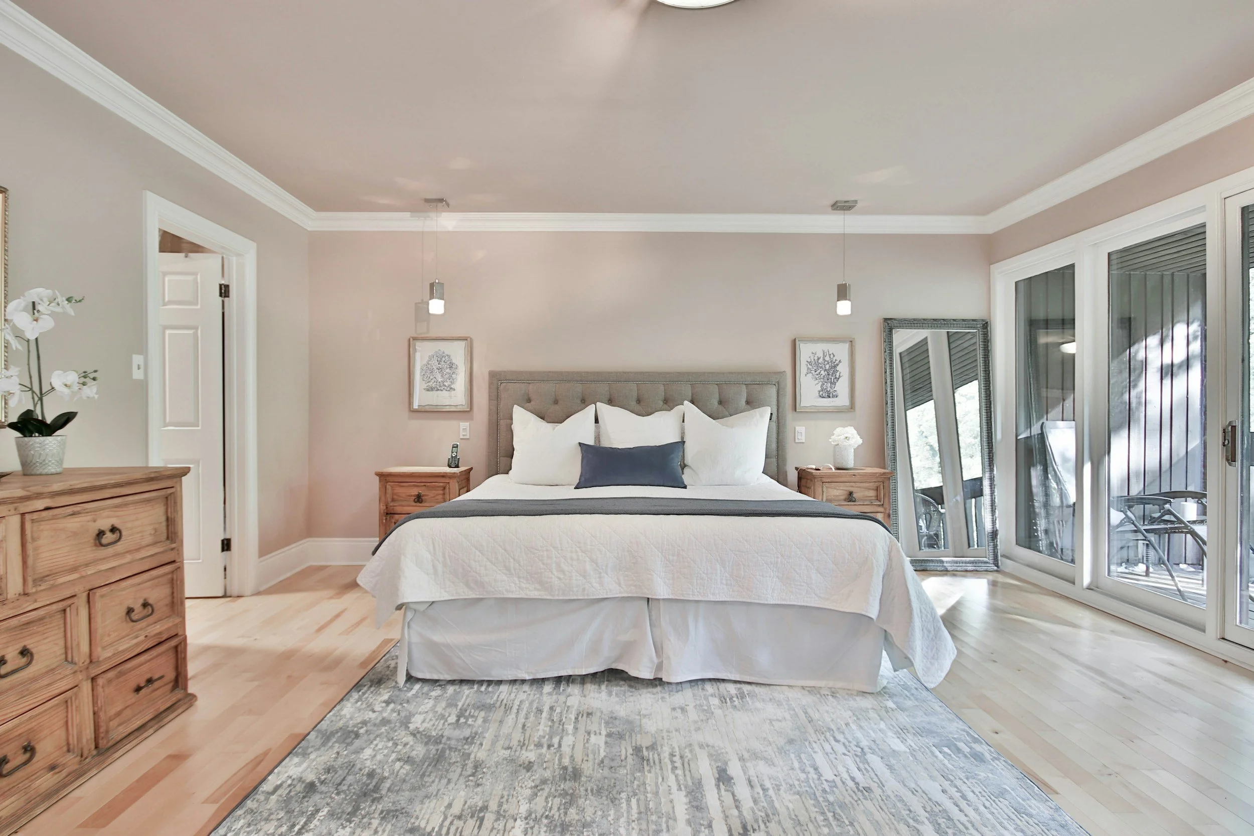 A bedroom with a queen-sized bed, using white bedding and decorative pillows, flanked by two wooden nightstands with lamps. There is a large mirror leaning against the right wall near sliding glass doors that lead to a balcony. The room has light-colored hardwood floors, a patterned area rug, and framed artwork on the wall above the bed.