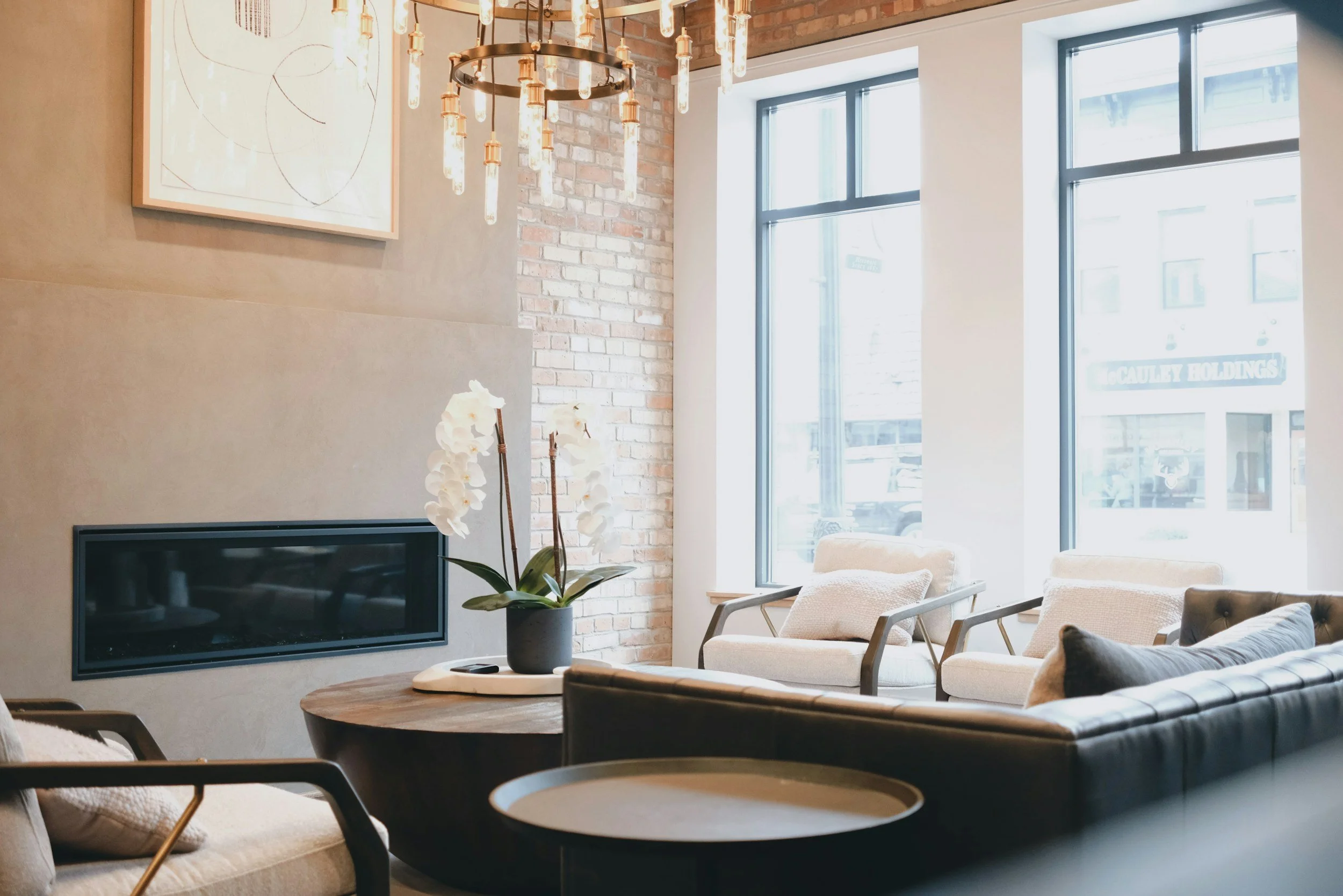 Cozy modern living room with beige and black furniture, a round wooden coffee table with an orchid plant, large windows, brick wall, and abstract artwork.
