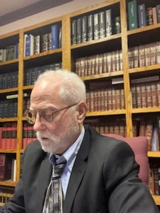 An elderly man with glasses, white hair, and a beard in a suit sitting in front of bookshelves filled with legal books or reference volumes.