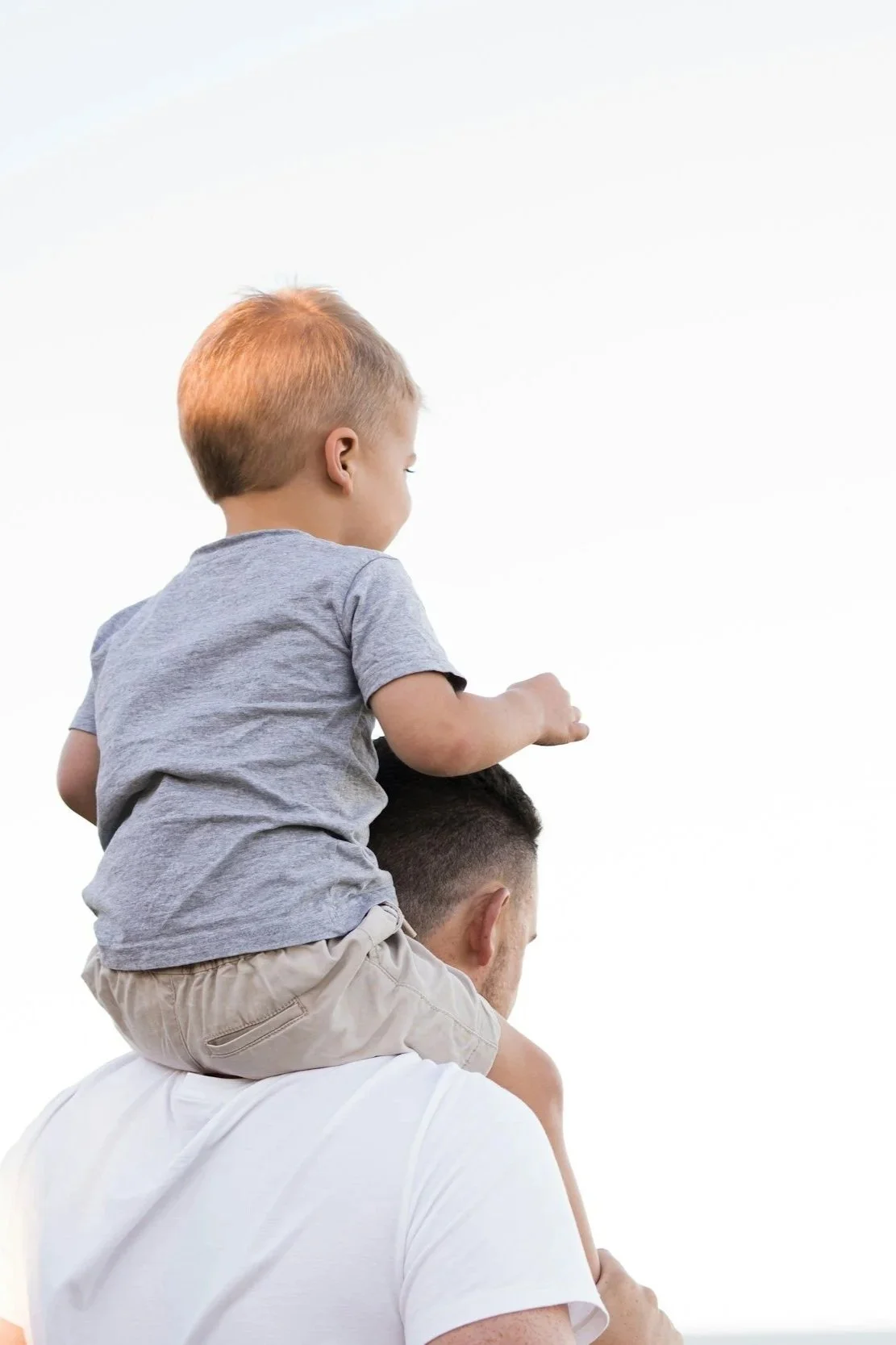 A young boy sitting on an adult man's shoulders, wearing a gray shirt and beige shorts, with a plain white background.