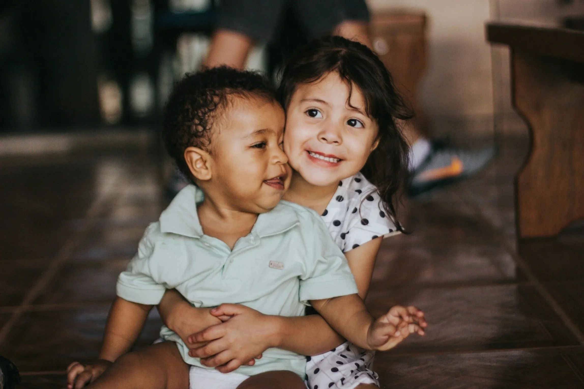 A young girl hugging a toddler boy from behind on a wooden floor, smiling, with furniture in the background.