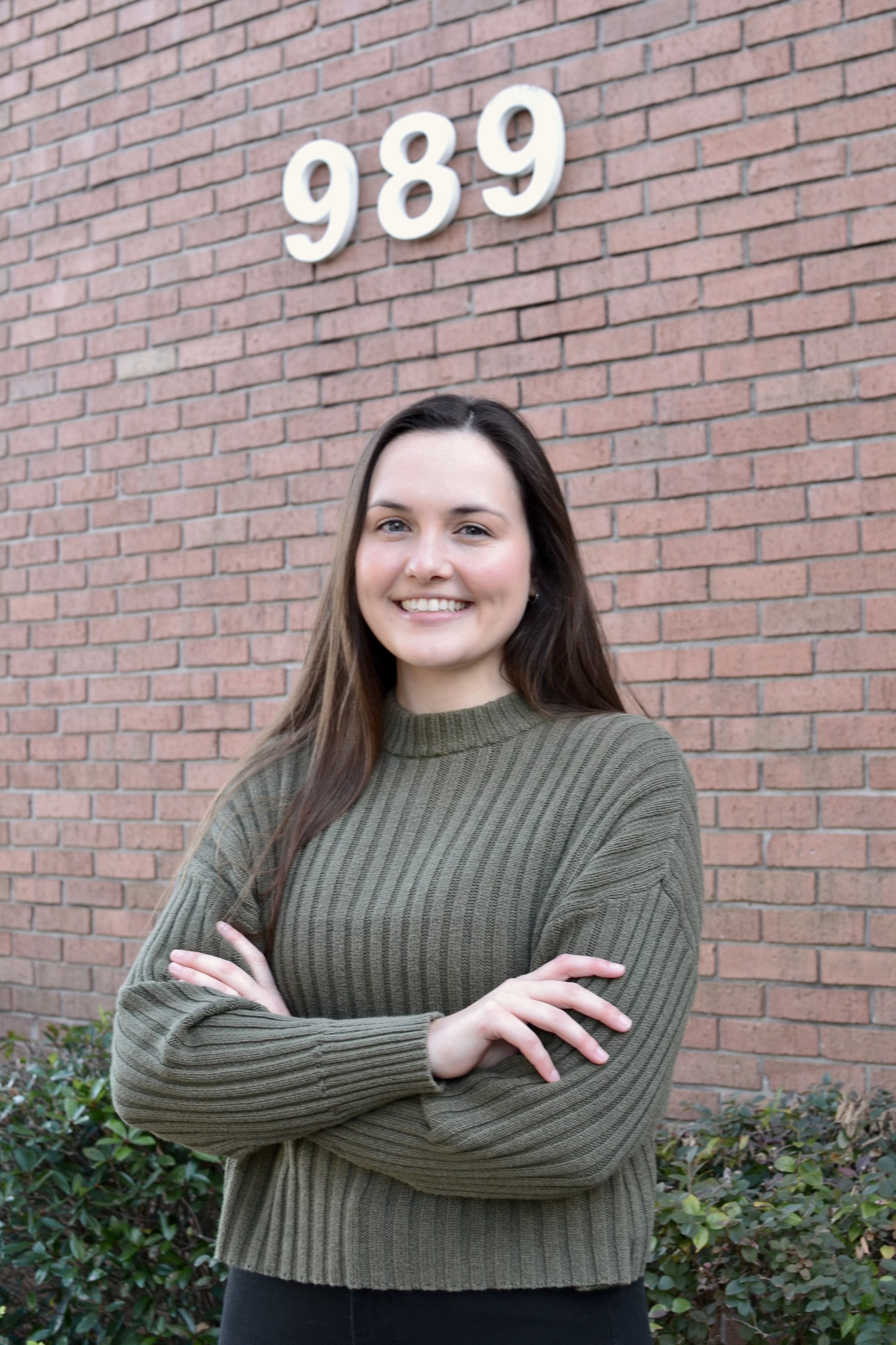 A woman with long brown hair, wearing a green ribbed sweater, crossed arms, standing outdoors in front of a brick wall with the numbers 989 mounted on it, and some green shrubbery at the base.