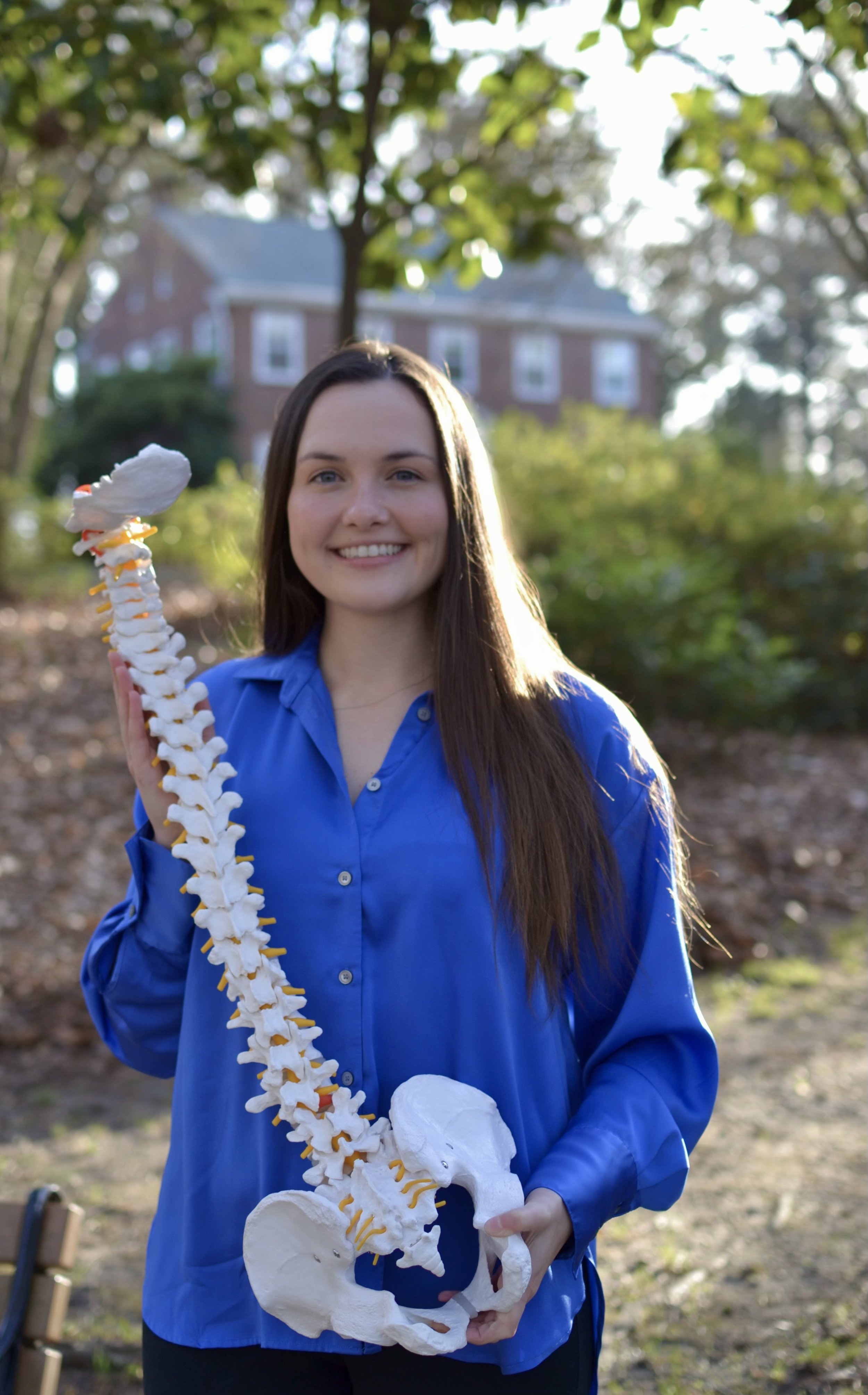 A young woman with long brown hair smiling, wearing a blue shirt, holding a model of a human spine in an outdoor setting with trees and a house in the background.
