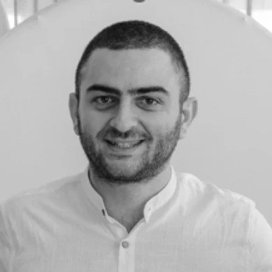 A man with short dark hair and a beard, smiling, wearing a white shirt, posed in front of a plain background.
