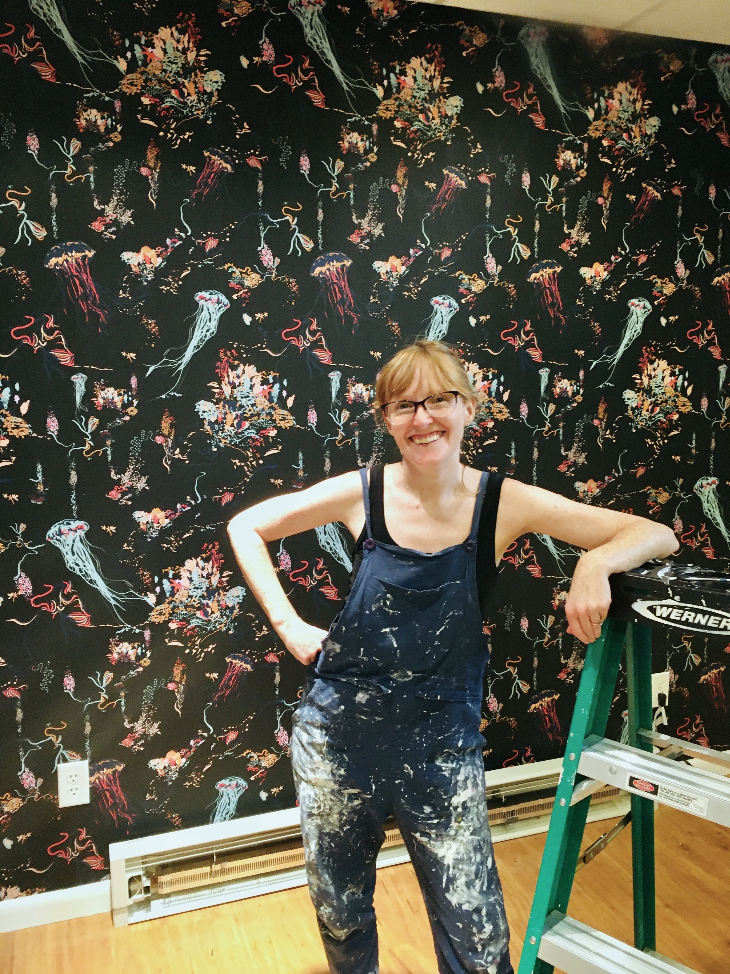 Tricia Wallace leaning on a ladder in paint covered overalls. She stands in front of colorful wallpaper featuring mint, red, and yellow jellyfish & sea flora on a black background