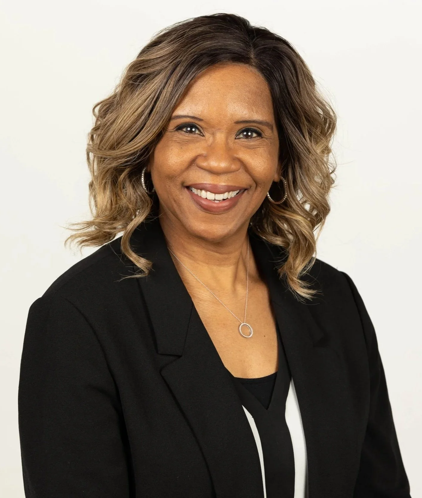 A woman with shoulder-length wavy hair, wearing a black blazer and a necklace, smiling against a plain white background.