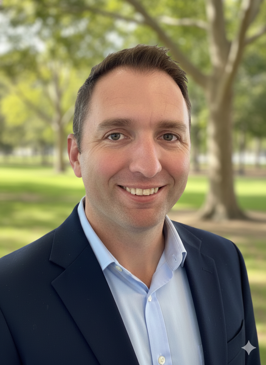 Portrait of a man in a suit smiling in a park with green trees and sunlight in the background.