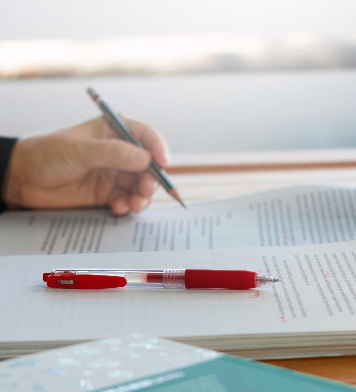 A person holding a pencil over an open book with printed text, with a red pen resting on the book.