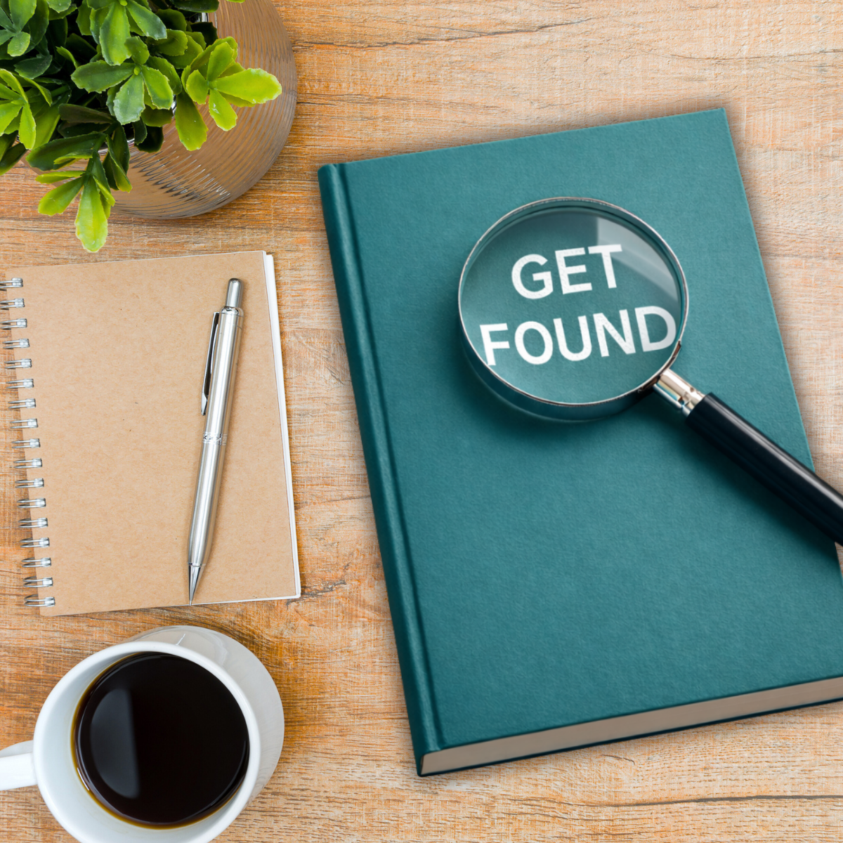Desk with a green plant, notebook with a pen, a cup of coffee, and a book with a magnifying glass on top showing the words "GET FOUND".