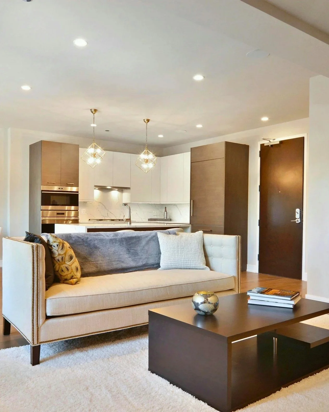 Sala moderna con sillón beige, mesa de centro negra, y cocina abierta con gabinetes blancos y oscuros, iluminación empotrada y lámparas decorativas de oro con bombillas expuestas.