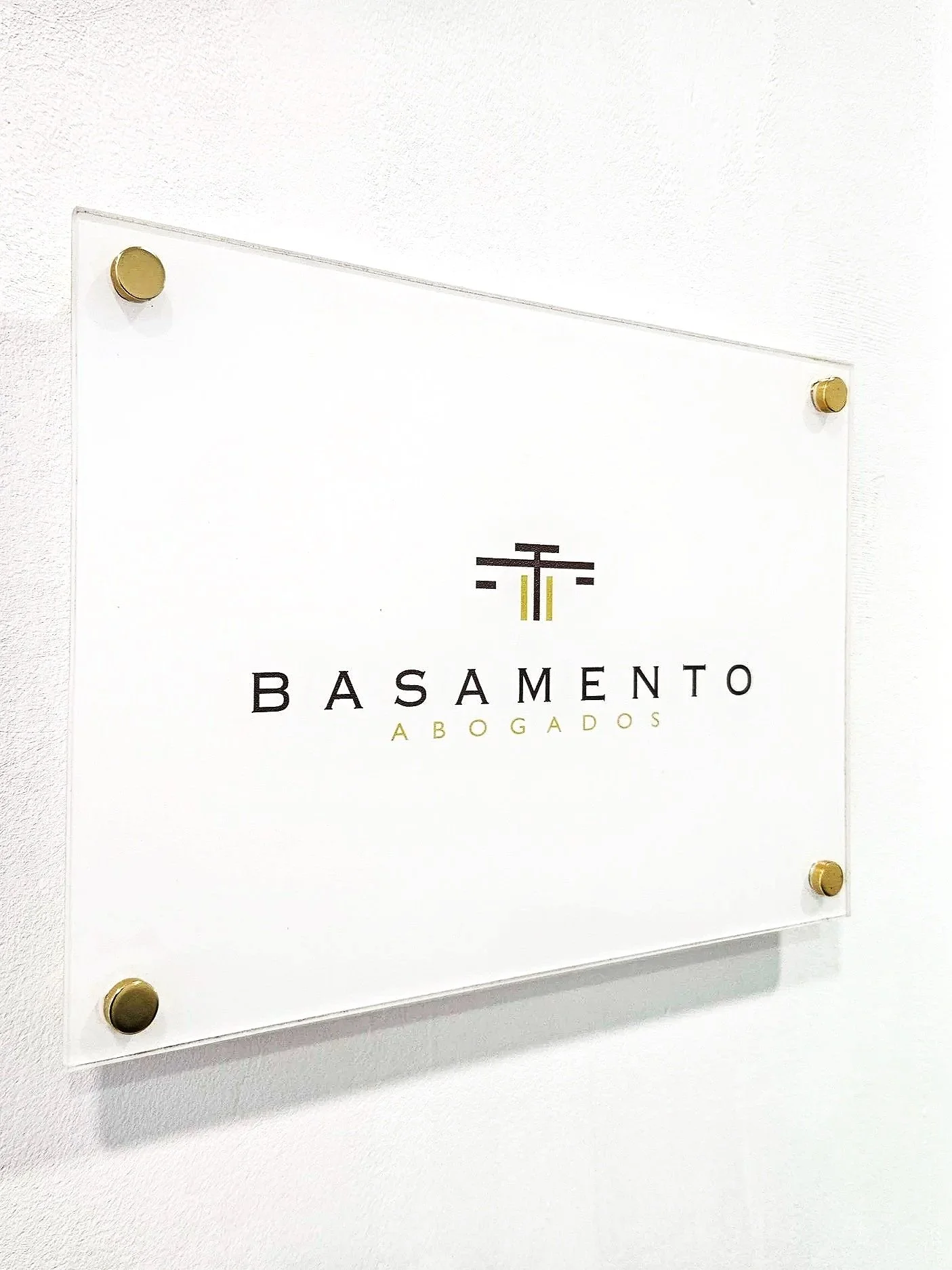Cartel de vidrio con logotipo y texto que dice 'Basamento Abogados' en una pared blanca.