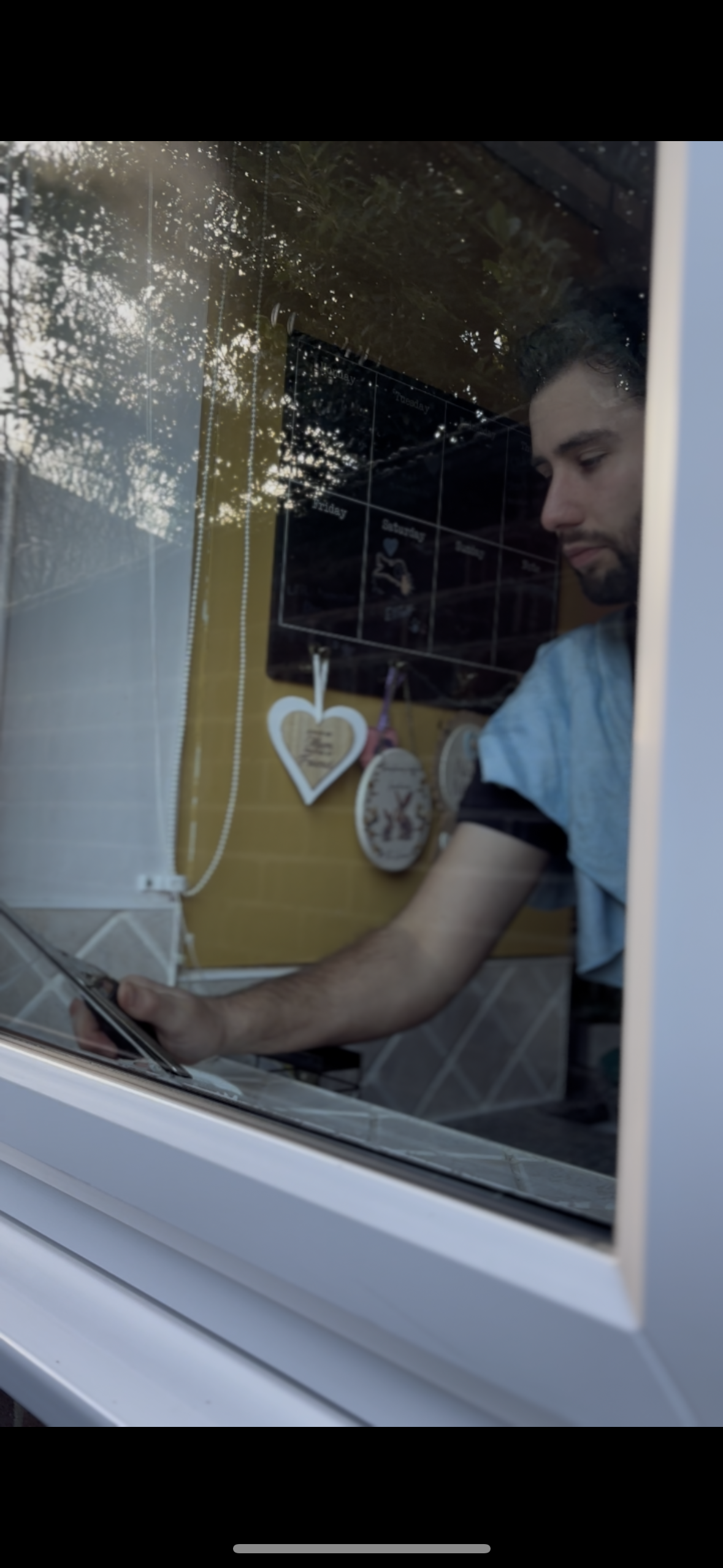 A man looking at his tablet through a window, with a reflection of trees and sky, inside a room decorated with wall hangings and a calendar.