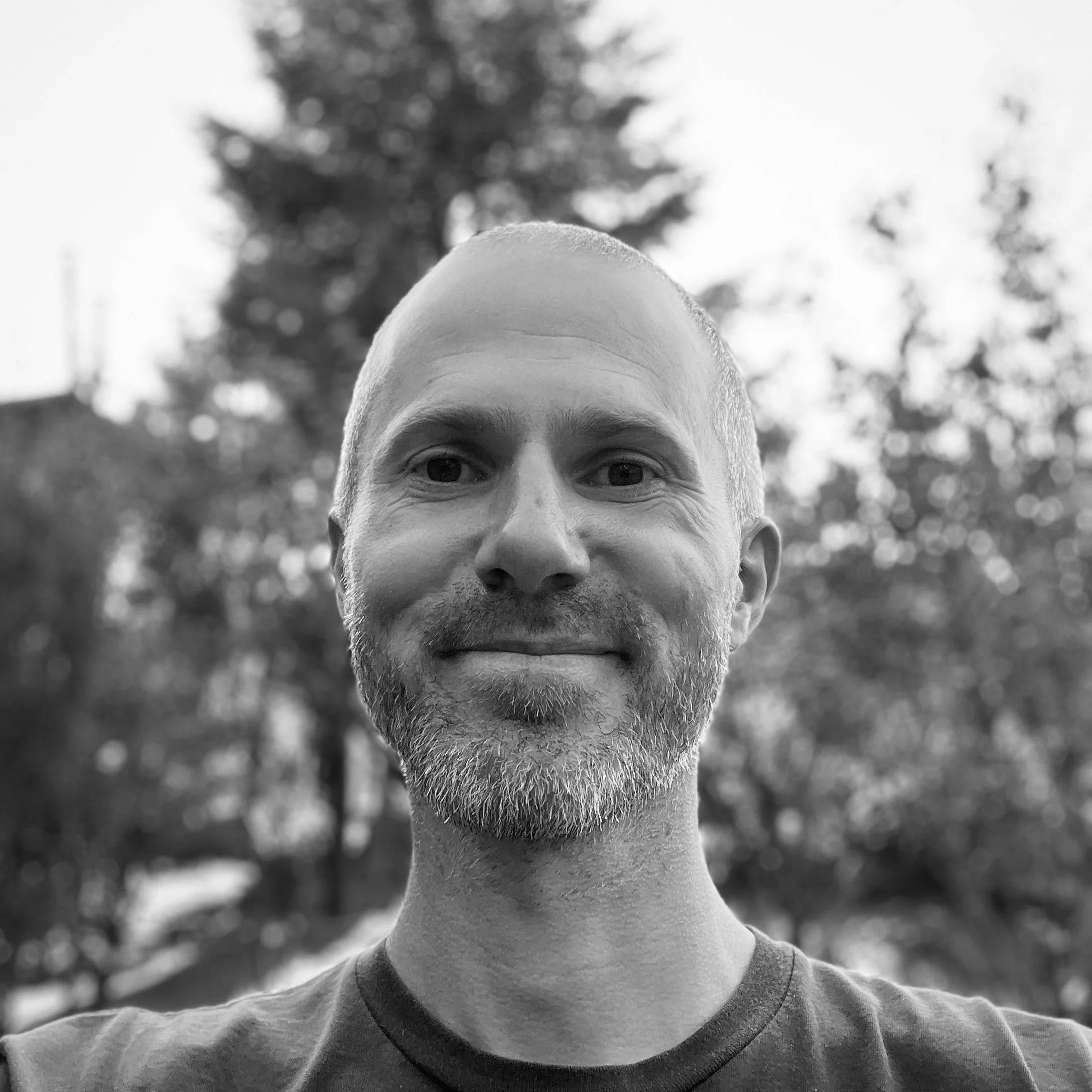 Black and white photo of a man with a beard and short hair outdoors, smiling at the camera with trees in the background.