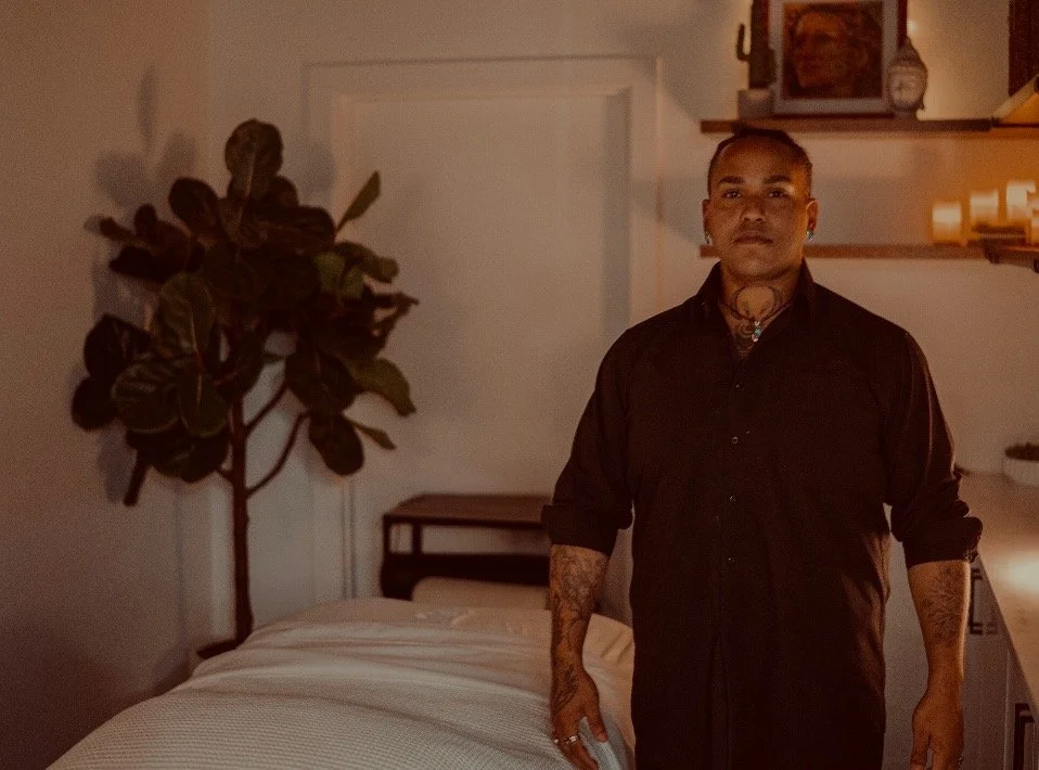 A man with tattoos standing in a bedroom with a bed, a potted plant, and shelves with candles and picture frames.