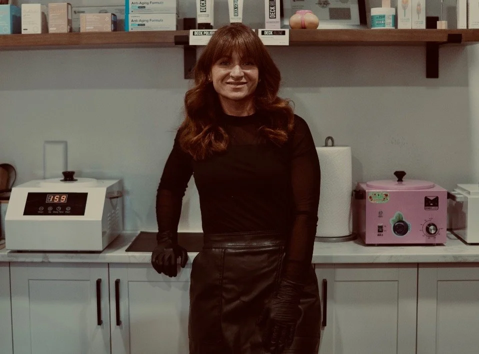 Woman with long reddish hair wearing black gloves and a black outfit standing in a laboratory or skincare clinic, with shelves of boxes and containers behind her and laboratory equipment on the counter.