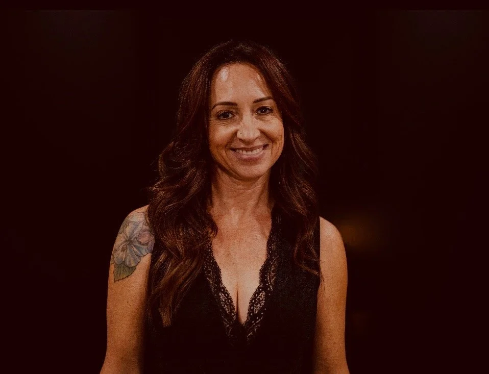 A woman with long brown hair, wearing a black lace top, smiling, with a tattoo on her left shoulder, against a dark background.