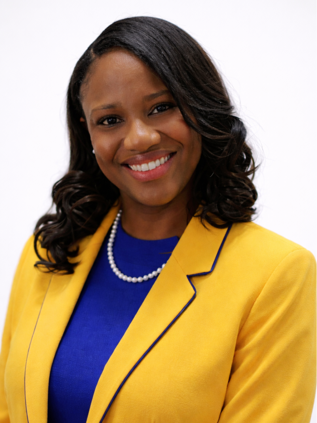 Portrait of a smiling Black woman with dark, styled hair, wearing a yellow blazer over a blue top, a pearl necklace, and earrings, against a plain white background.