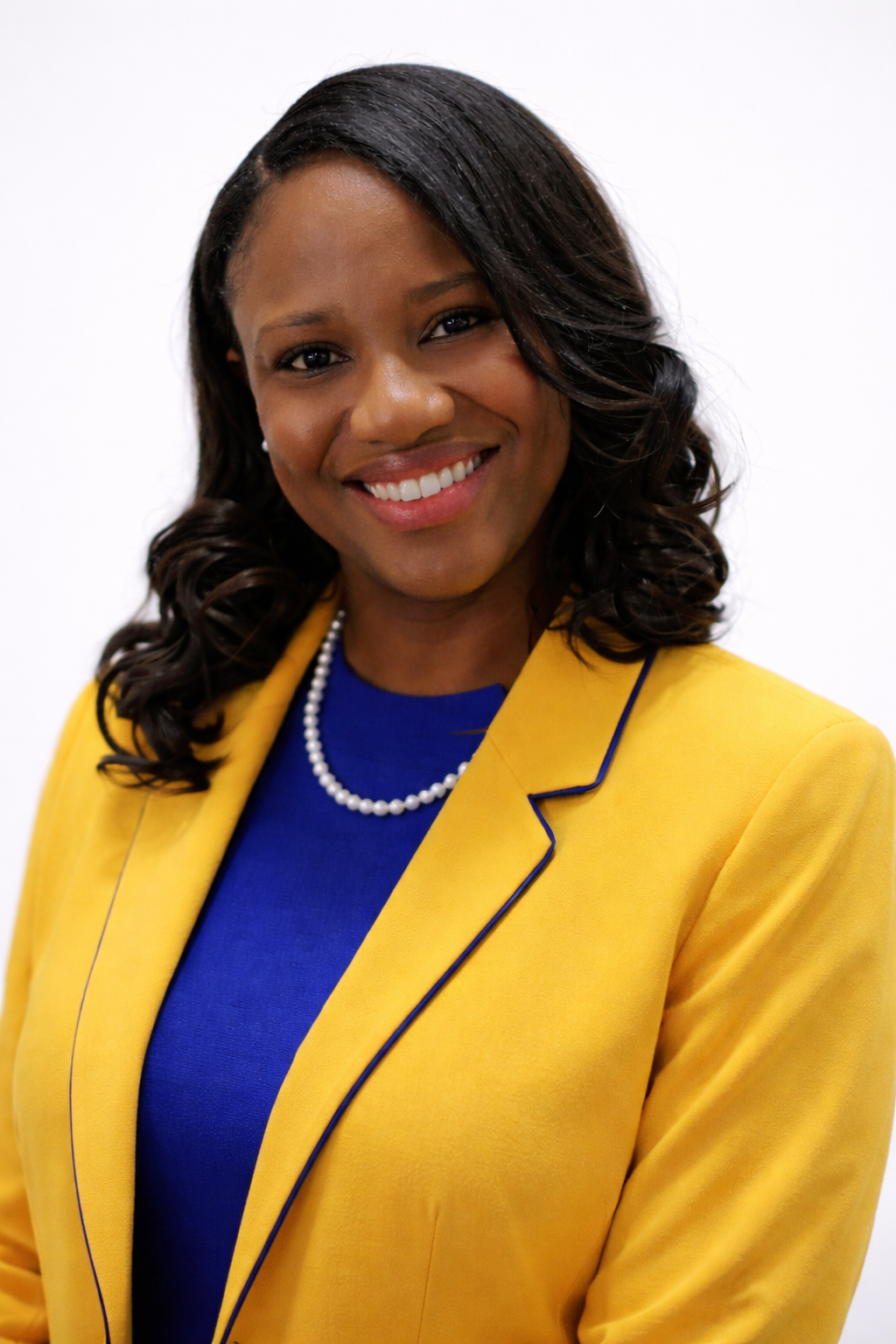 A smiling African American woman with shoulder-length curly hair, wearing a yellow blazer with blue piping, a blue top, and a pearl necklace, against a white background.