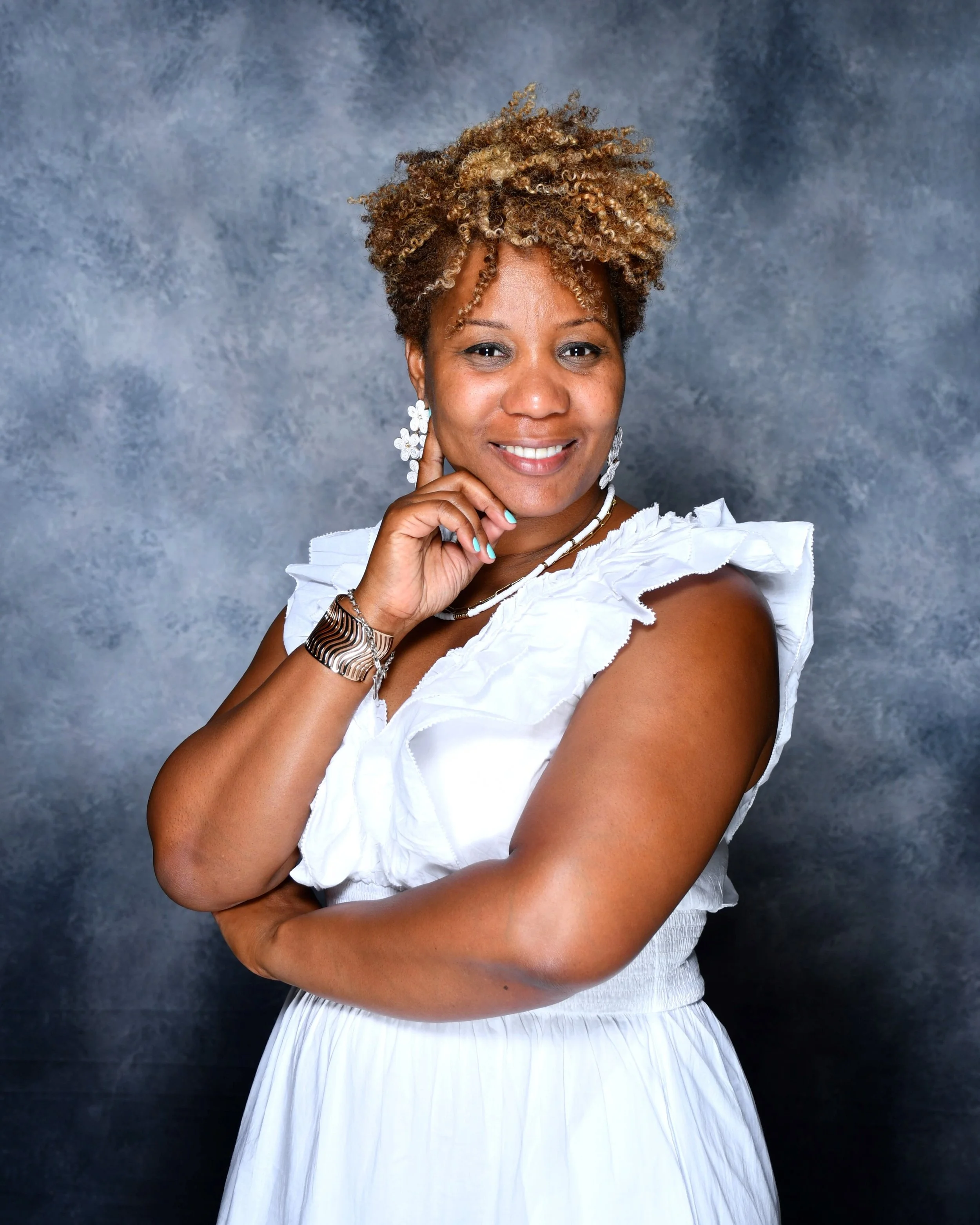 A woman with curly, blonde-tinted hair is smiling, resting her chin on her right hand, wearing a white ruffled dress and jewelry, standing against a textured gray background.