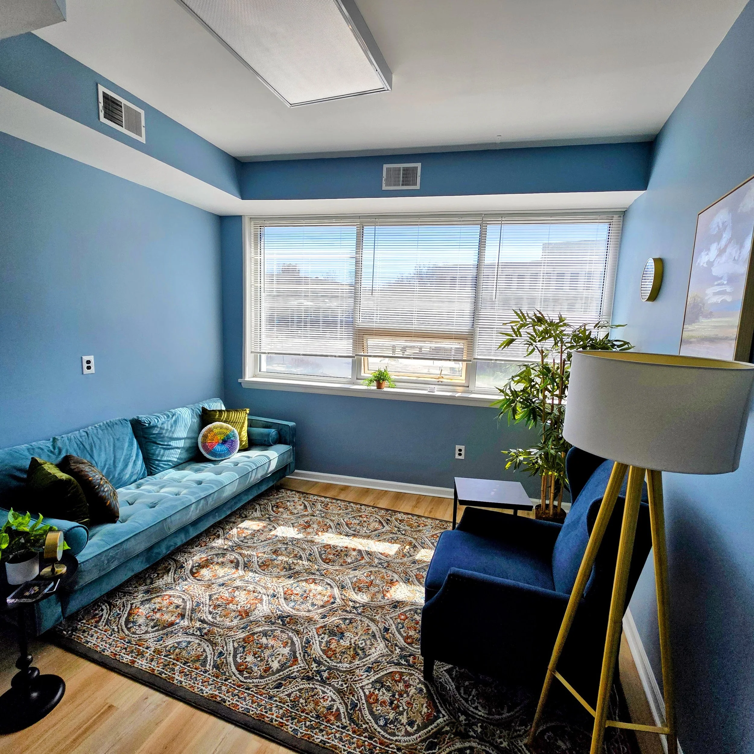 A living room with blue walls, a large window with blinds, a turquoise sofa with pillows, a patterned rug, a black armchair, a tall yellow floor lamp, a large plant, and framed artwork on the wall.