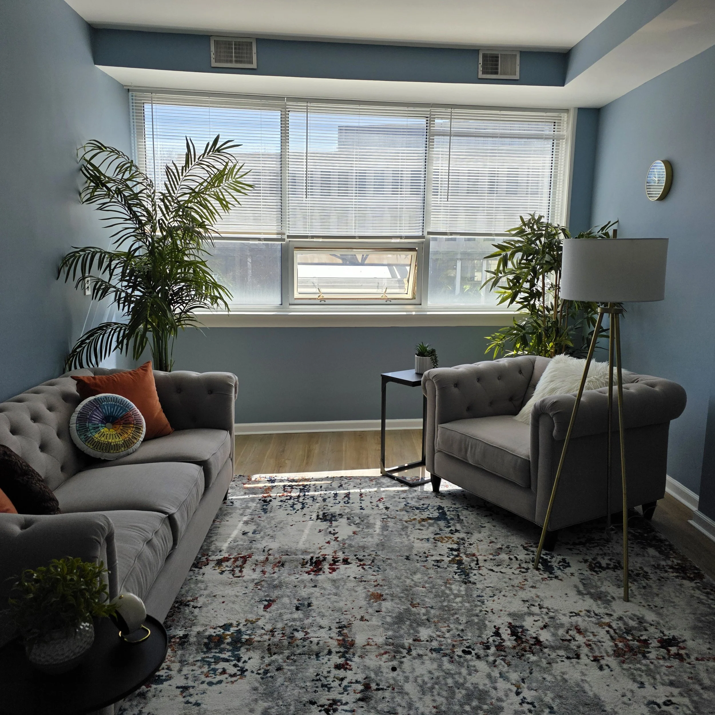 A modern living room with a large window, two gray tufted sofas, a lamp, plants, and a colorful area rug.