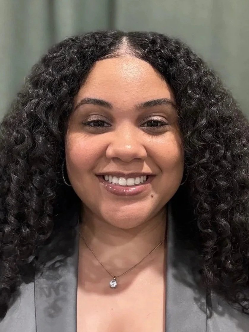 A woman with curly black hair smiling, wearing a gray blazer, a necklace with a large pendant, and hoop earrings.