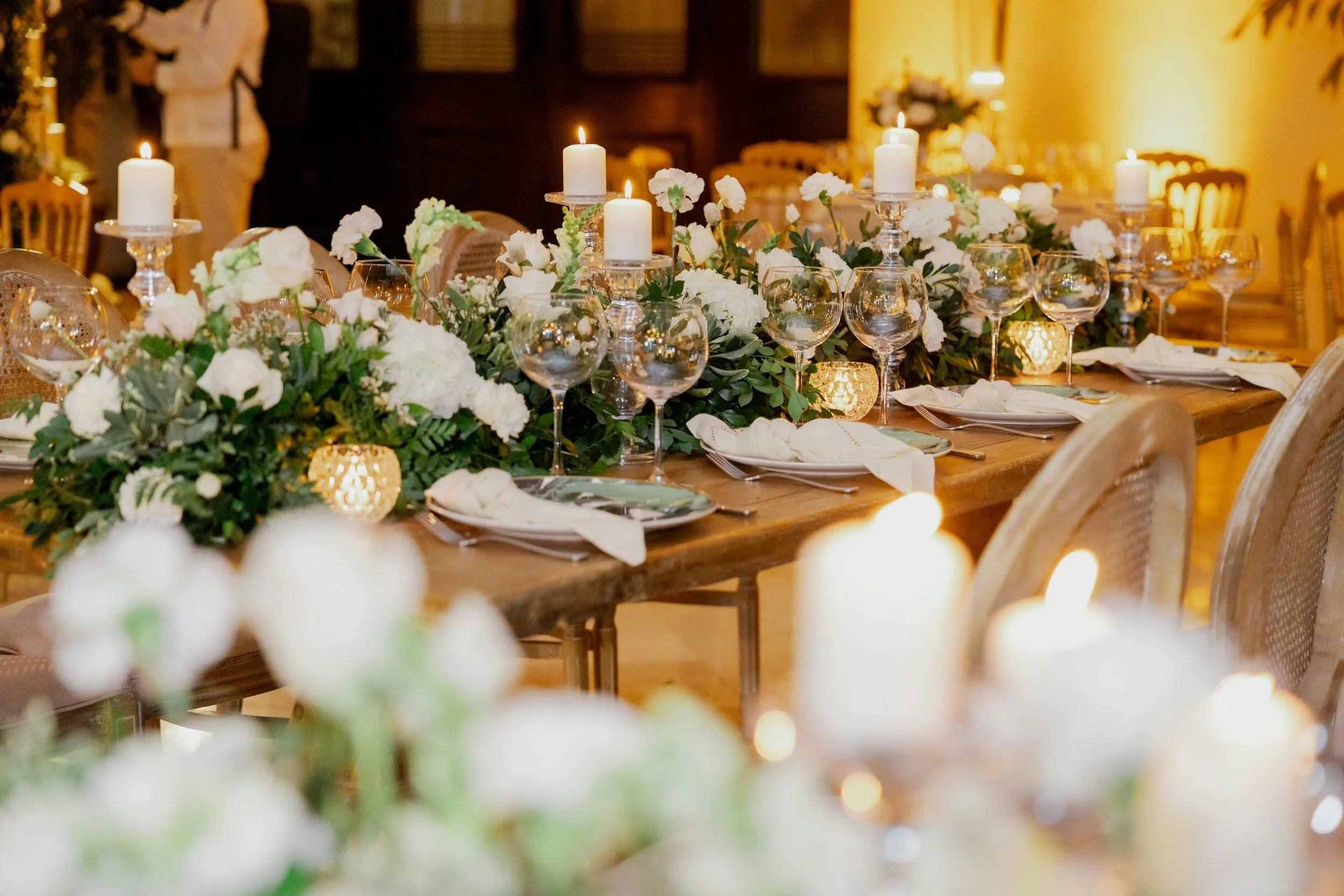 Elegant dining table decorated with white flowers, green foliage, lit candles, and glassware in a warmly lit event setting.