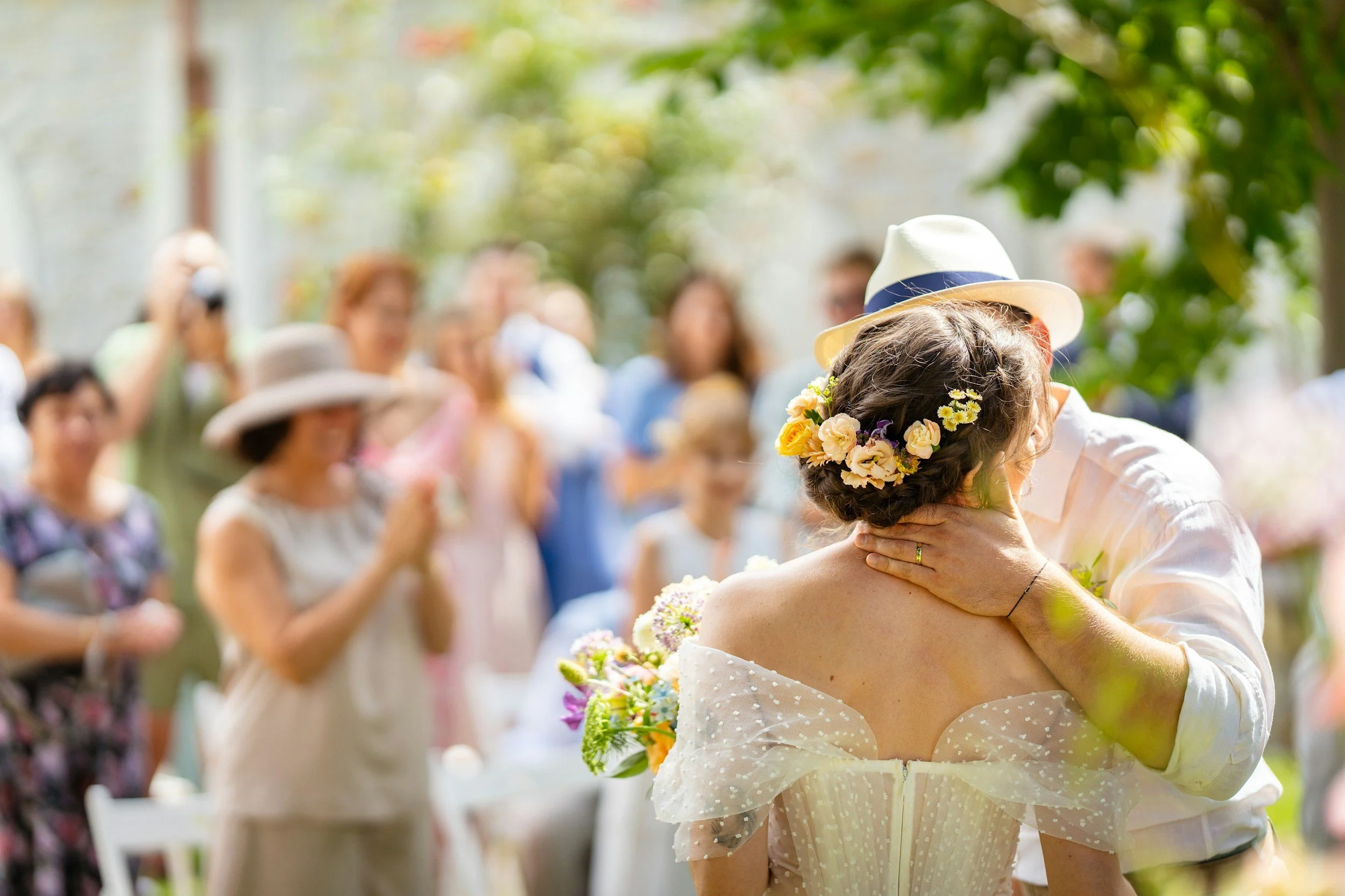 A couple is sharing a kiss at a wedding reception outdoors, surrounded by celebrate guests and lush greenery.