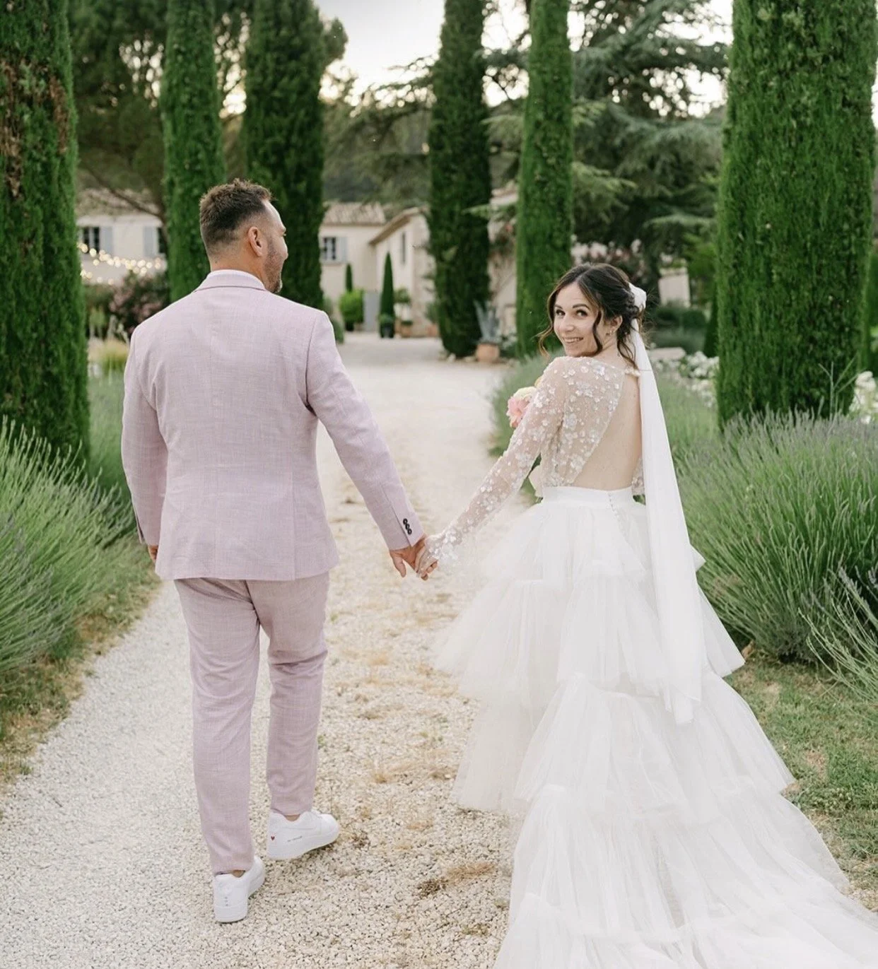 A bride and groom walking hand in hand along a garden path, with the bride looking back and smiling at the camera. The bride wears a wedding dress with lace sleeves and a voluminous skirt, while the groom wears a light pink suit. Tall green cypress trees line the path.
