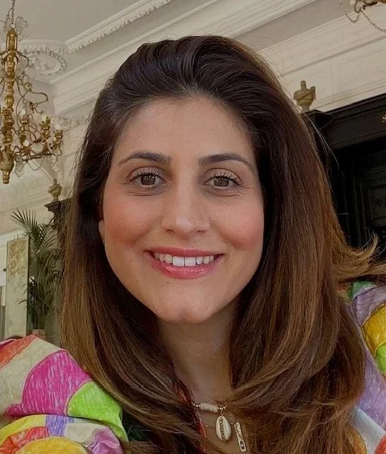 A woman with shoulder-length brown hair and light skin smiling indoors with ornate ceiling molding, gold chandelier, and a dark fireplace in the background.