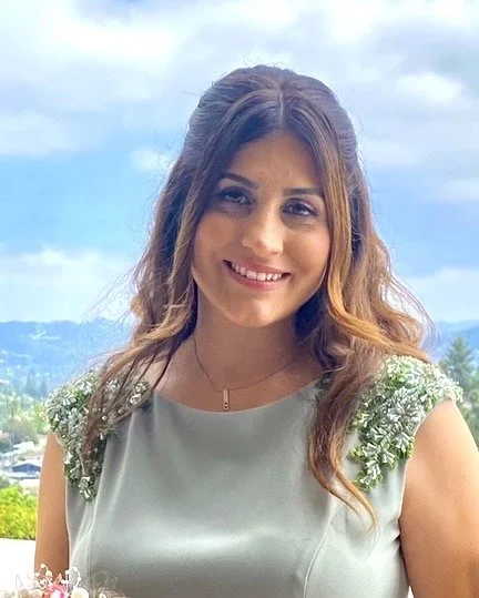 A young woman with long, wavy brown hair smiling outdoors, wearing a light-colored dress with floral shoulder details, against a background of a partly cloudy sky and distant mountains.