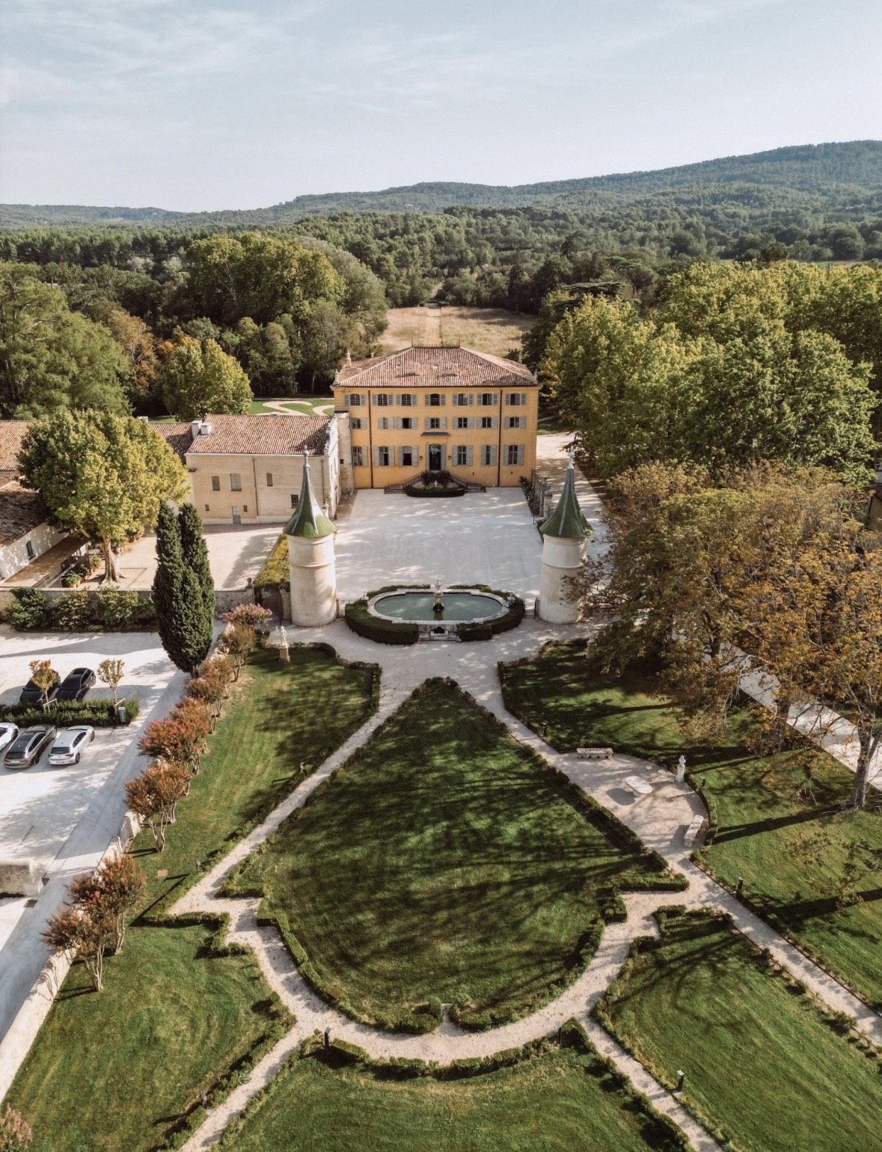 Aerial view of a large estate with a mansion, fountain, and manicured gardens surrounded by trees and hills.