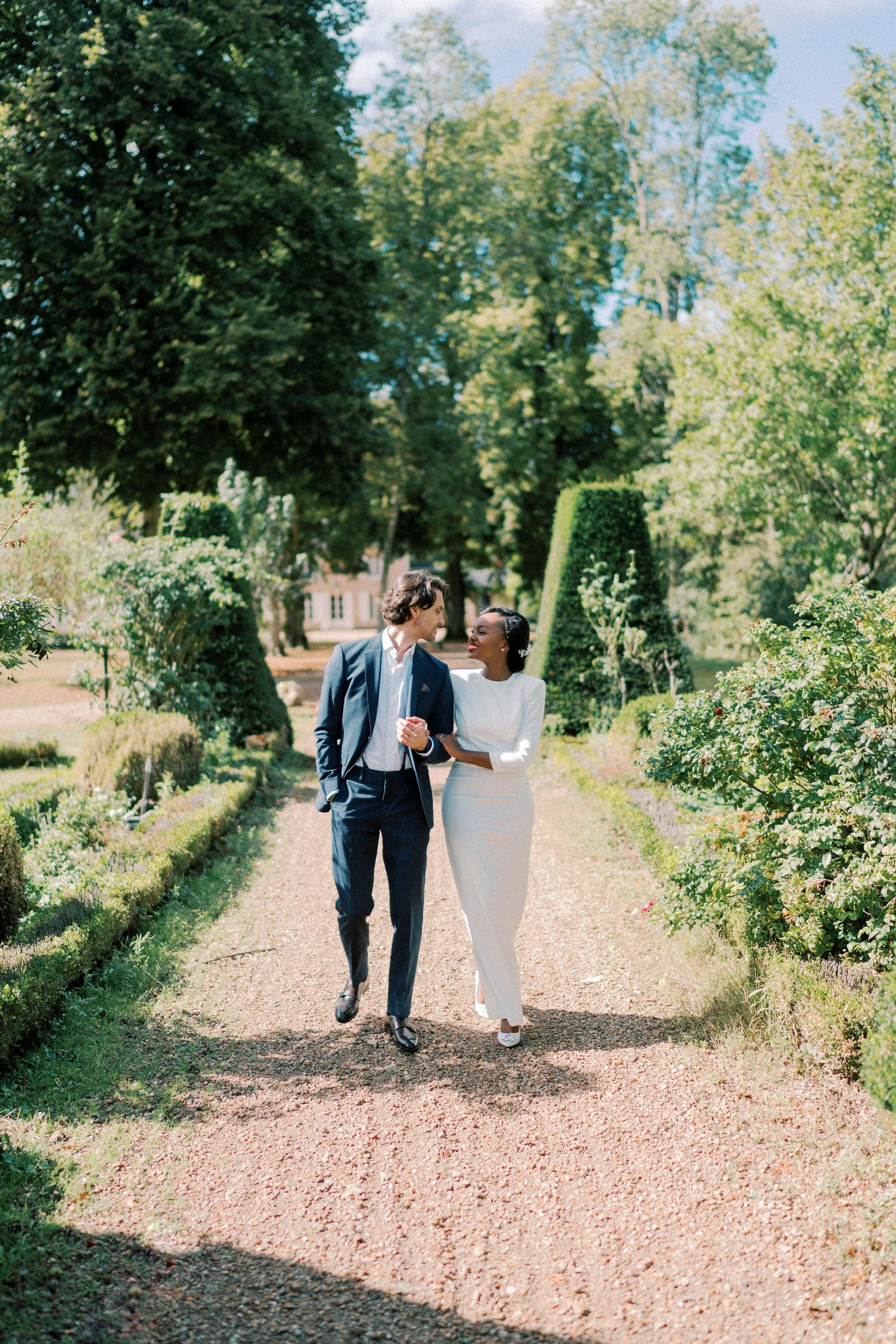 A man in a suit and a woman in a white dress walking together on a garden path, holding hands and smiling, surrounded by lush green trees and bushes on a sunny day.