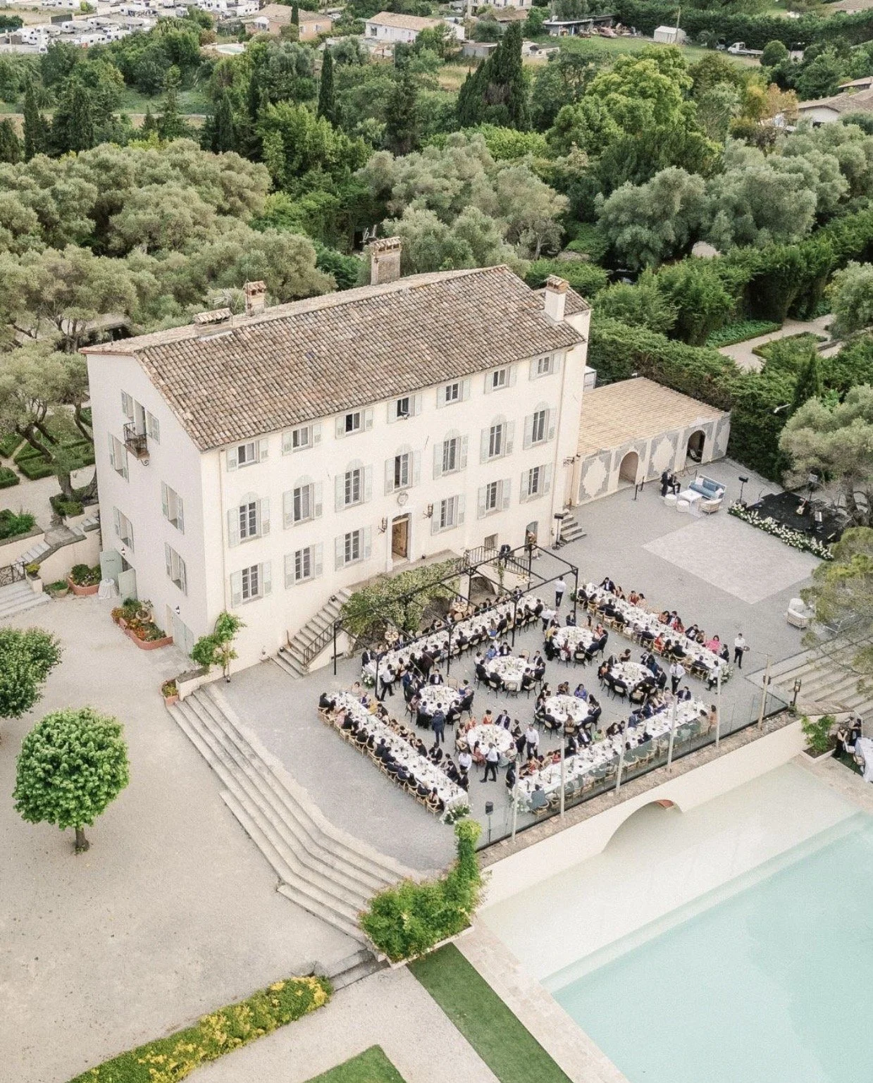 An outdoor event with guest tables set for a meal, next to a large, historic-looking building with a red-tiled roof, surrounded by lush green trees and landscaping.