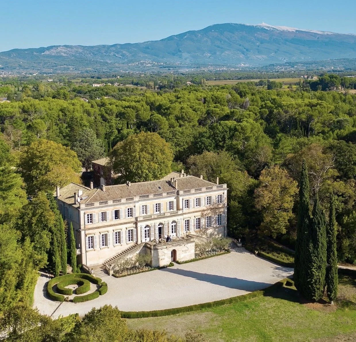 A large historic mansion with beige walls and tiled roof surrounded by manicured gardens and tall cypress trees, set in a lush green landscape with a mountain range in the background.