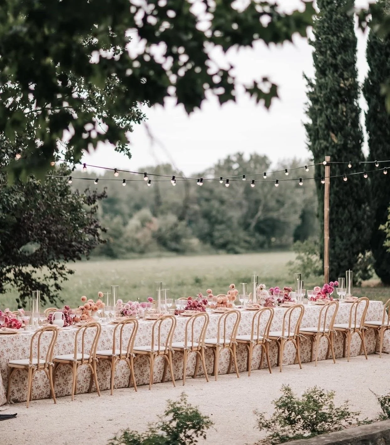 An outdoor dining setup with a long floral tablecloth-covered table, decorated with pink and peach flowers, candles, and glassware, under string lights on wooden poles in a garden setting with trees and greenery.