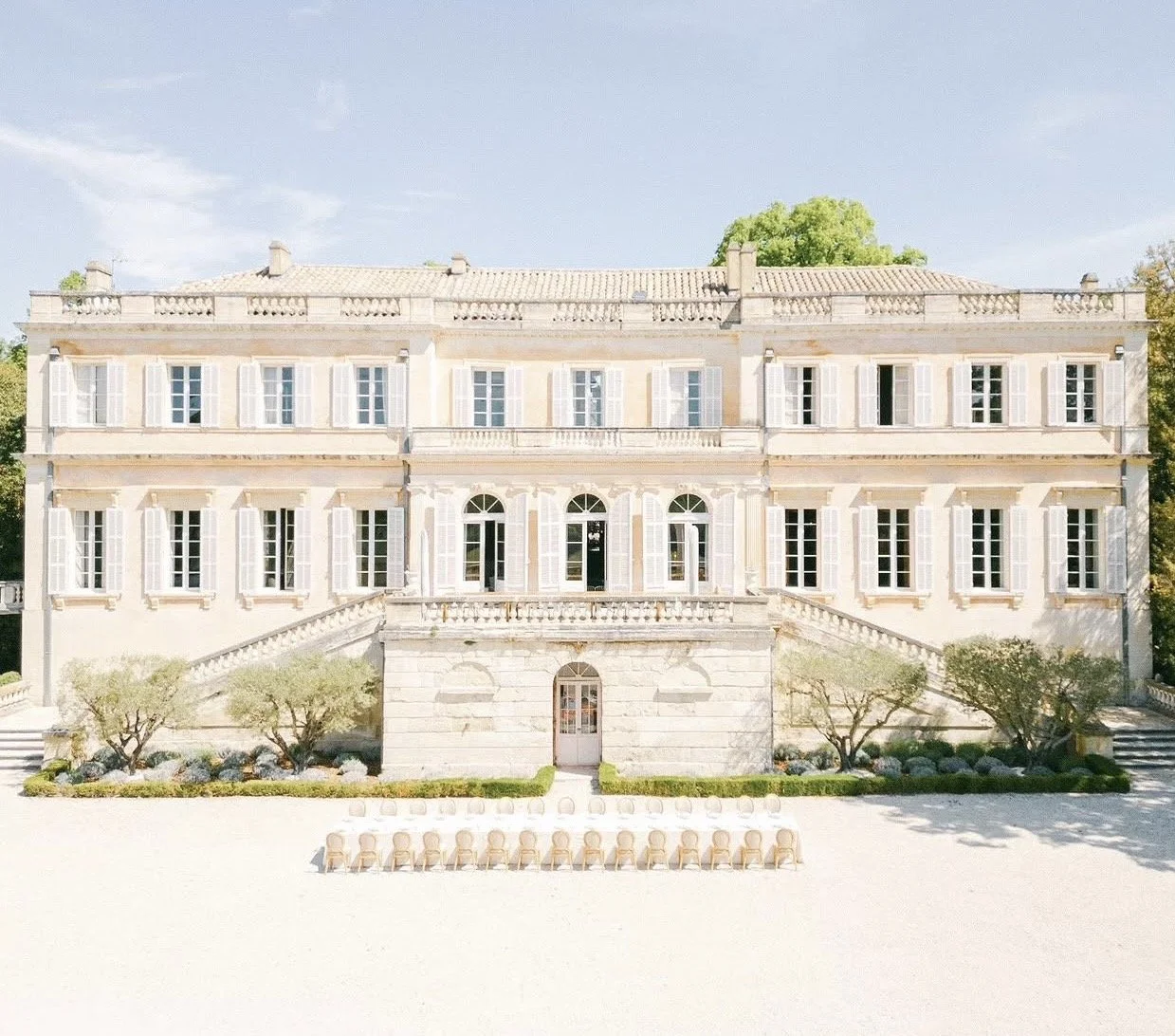 A large, elegant white mansion with multiple windows and stairs on both sides, set against a clear blue sky and green trees.