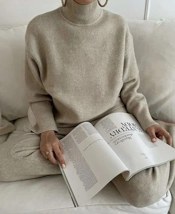 Person sitting on a white sofa, wearing a beige turtleneck sweater and matching pants, holding an open magazine.