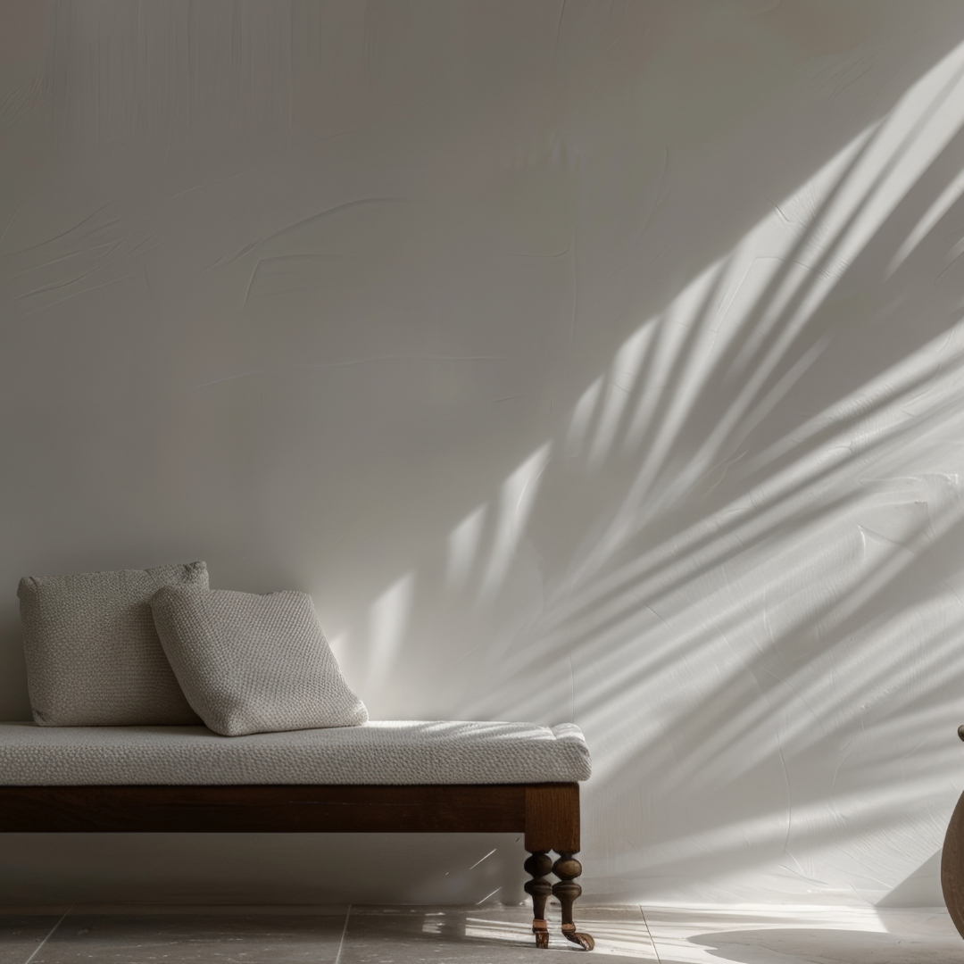 A minimalist room with a wooden daybed with white textured cushions, light-colored tiled floor, and a white wall with sunlight creating shadow patterns of a large plant.